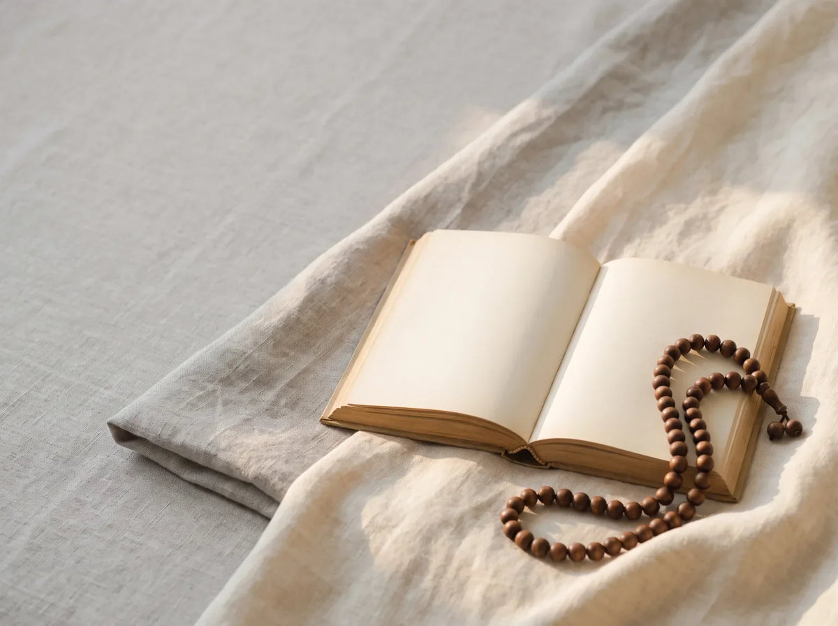 An open book on folded linen beside a strand of wooden prayer beads in soft morning light