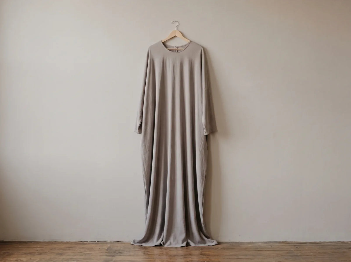 A loose one-piece prayer jilbab in dove grey hanging on a wooden hanger