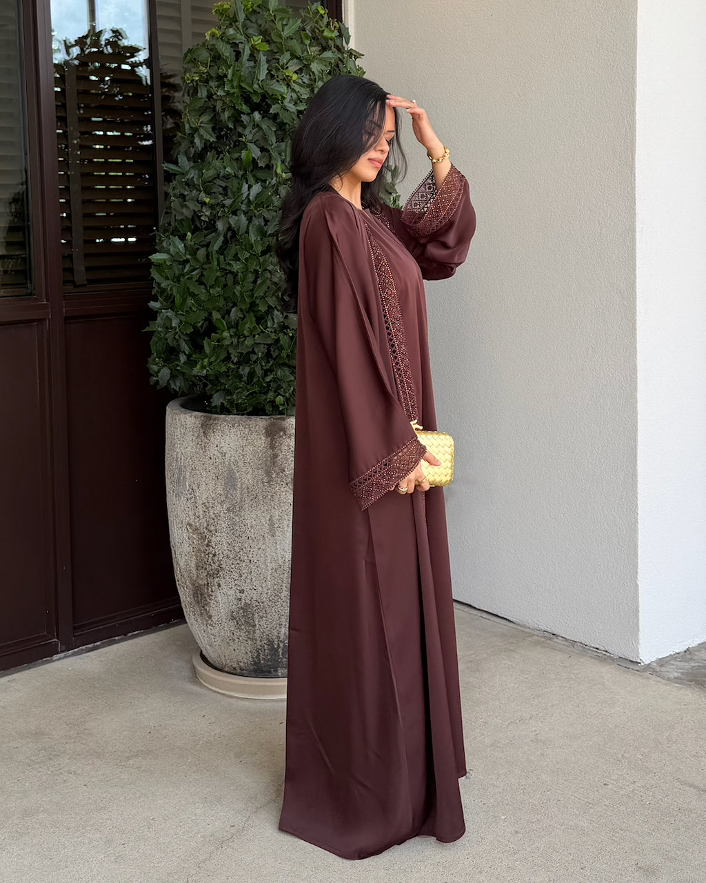 A woman wearing a long flowing brown abaya with lace trim beside a potted plant