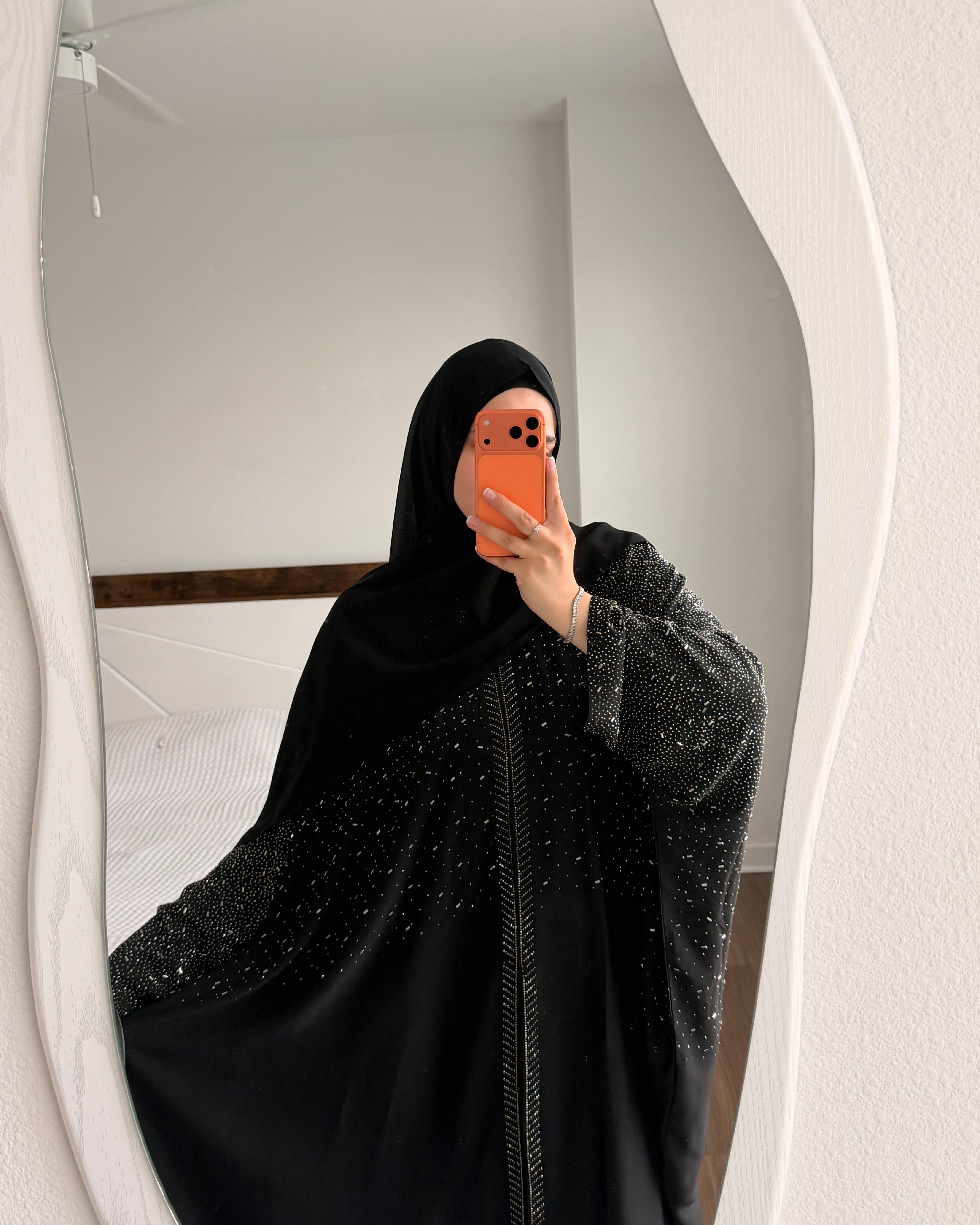 A muslim woman taking a mirror selfie wearing a black amina abaya with subtle silver glitter details and a matching black hijab, holding an orange phone.