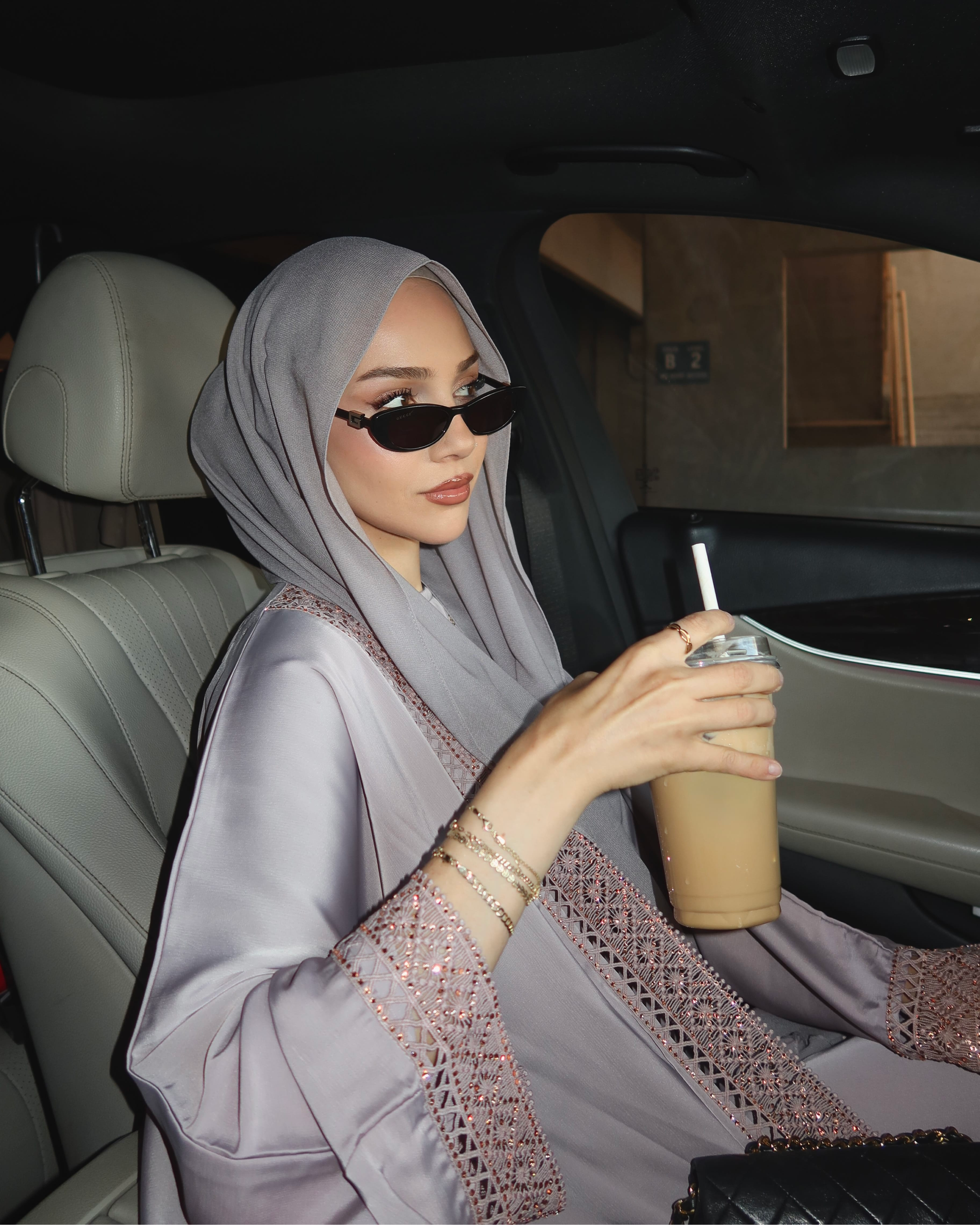 A woman wearing a light gray hijab and a matching gray abaya with embroidered details sits inside a car, holding an iced coffee.