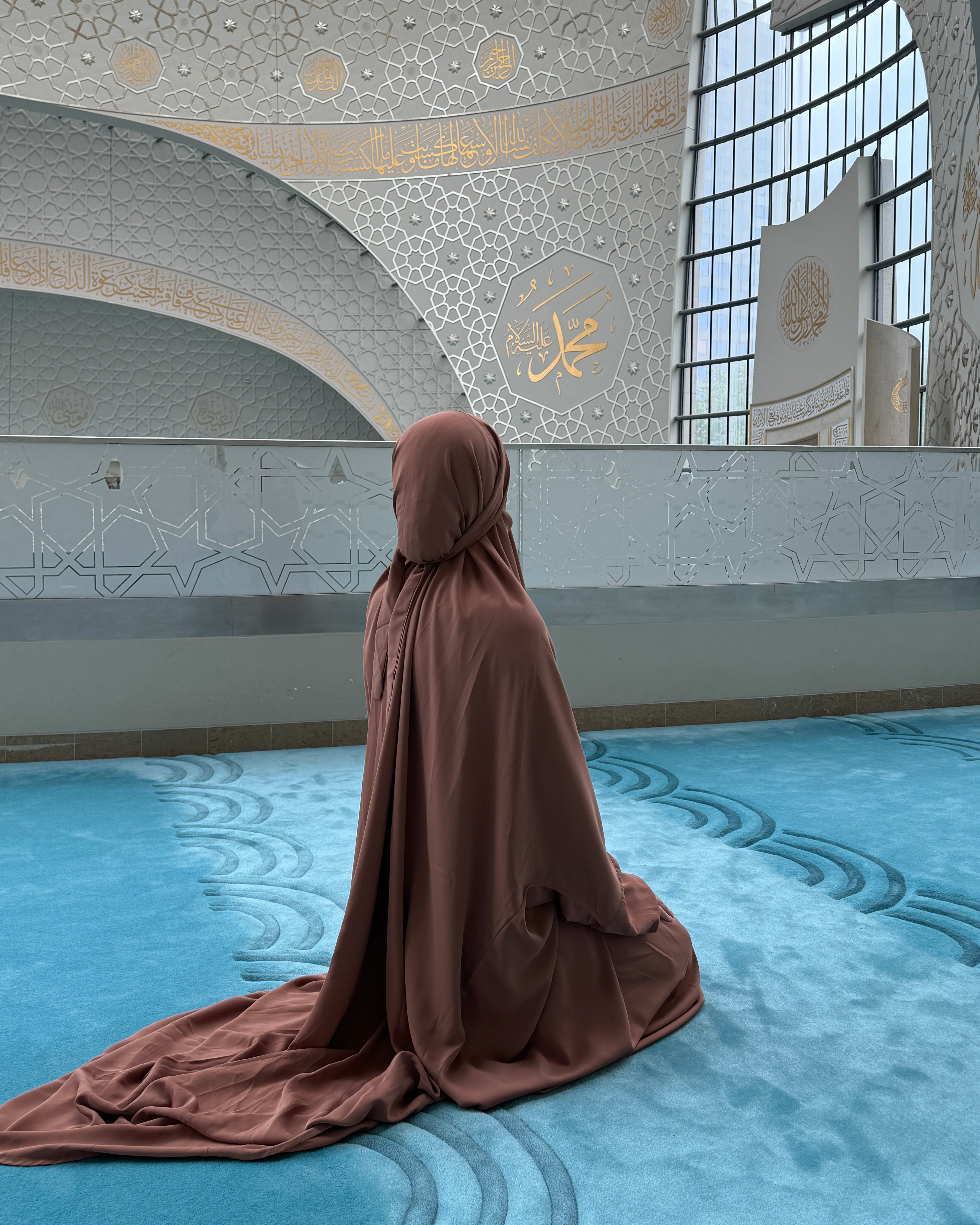 A person wearing a flowing brown modest garment and head covering kneels on a blue patterned prayer carpet inside a large mosque with arched ceilings, geometric wall details, and tall windows.
