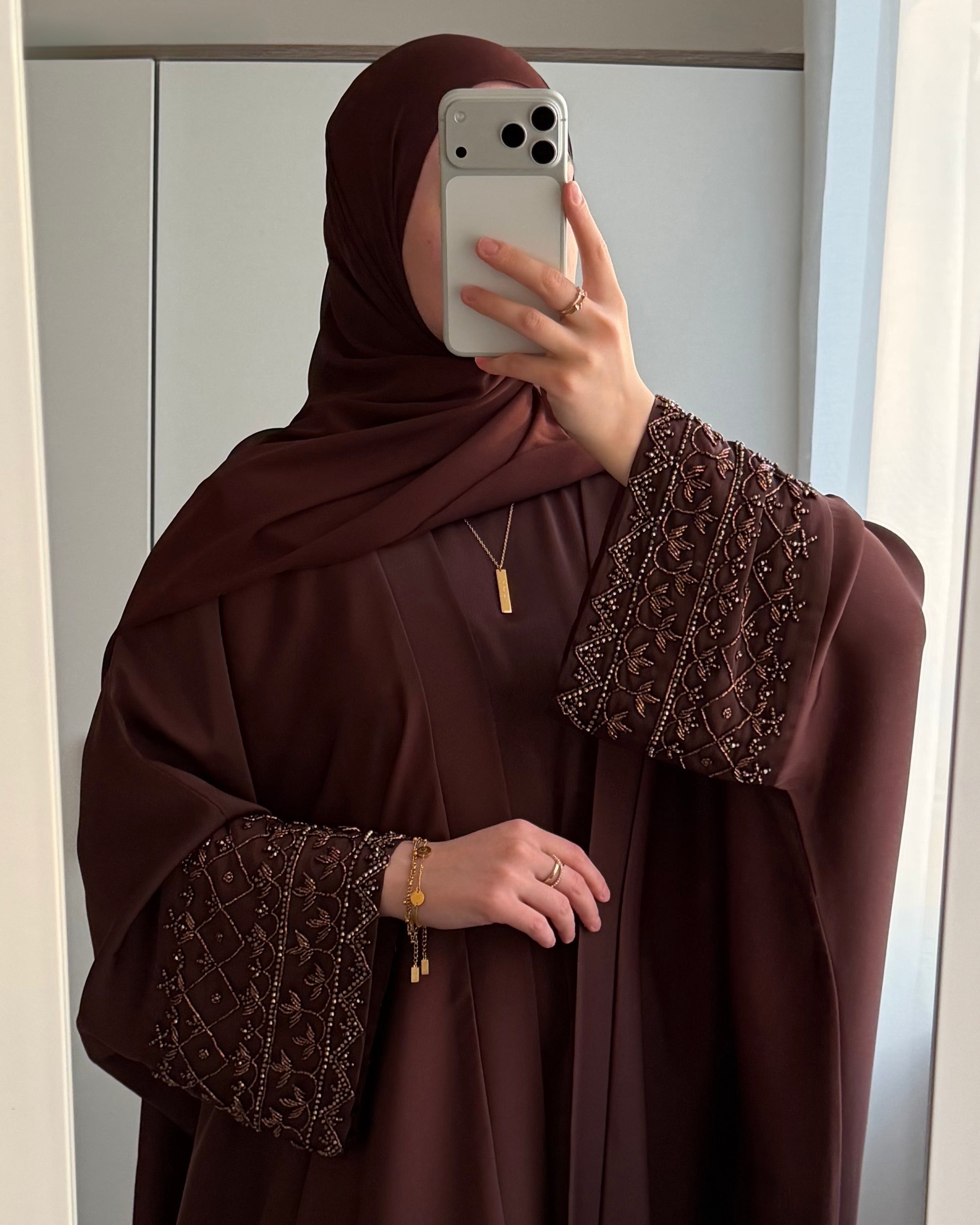 Muslim Woman wearing a brown abaya with intricate detailing, gold verily with hardship necklace and bracelet, taking a mirror selfie with her iphone.