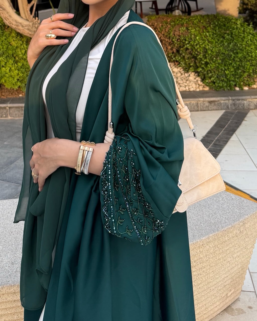 A woman wearing a flowing dark green abaya with wide sleeves and a matching headscarf, styled with a white inner top, a green beaded clutch, and a cream shoulder bag in an outdoor setting.