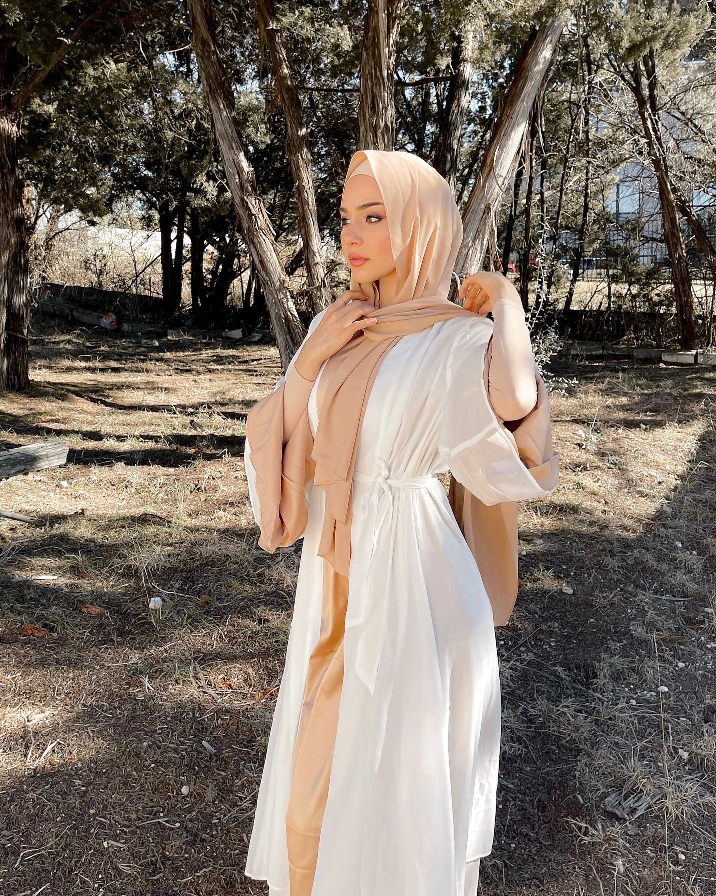 Woman in a white dress and beige hijab standing in a natural setting with trees.
