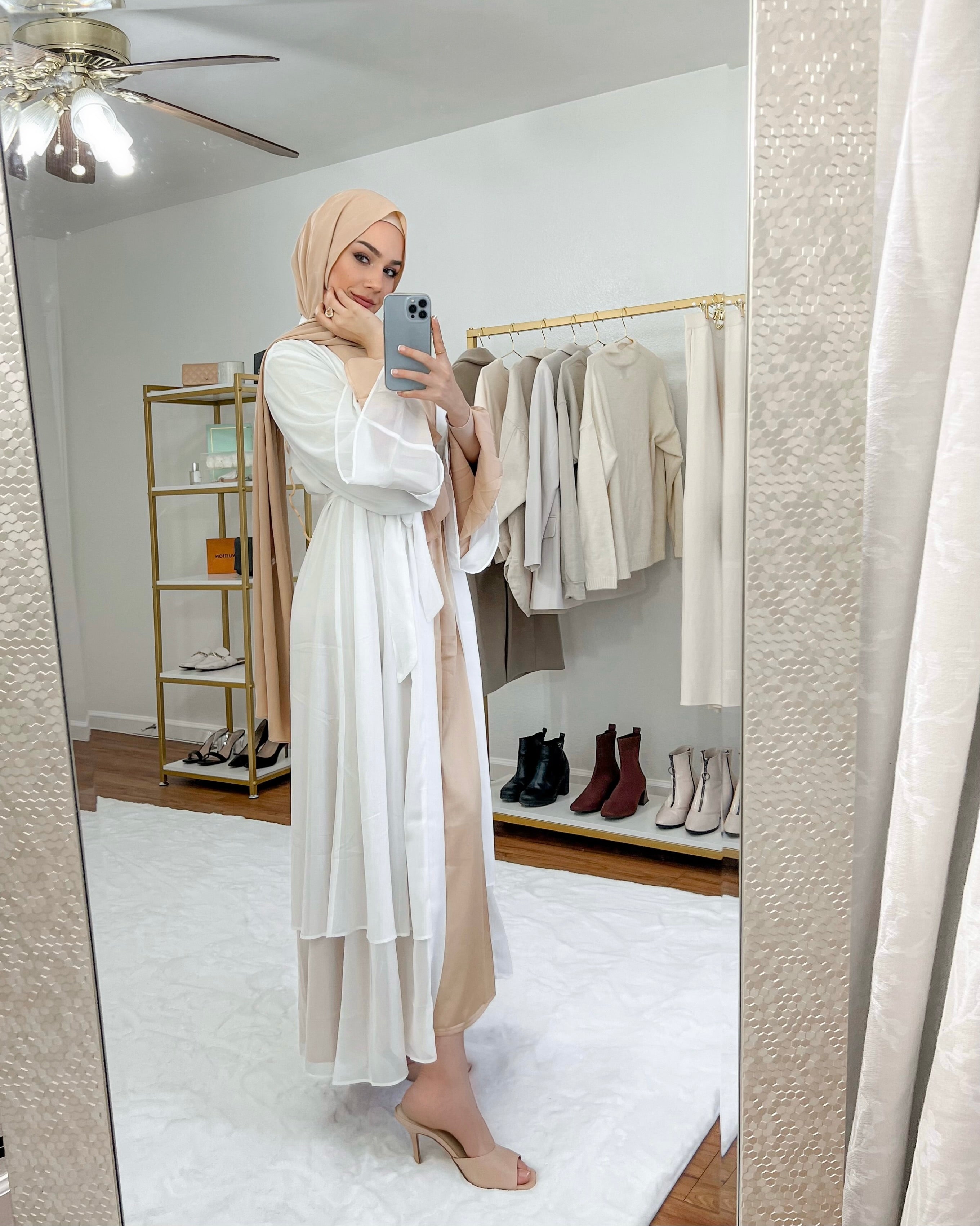 Woman in a white dress taking a mirror selfie in a room with clothing and shoes on shelves.