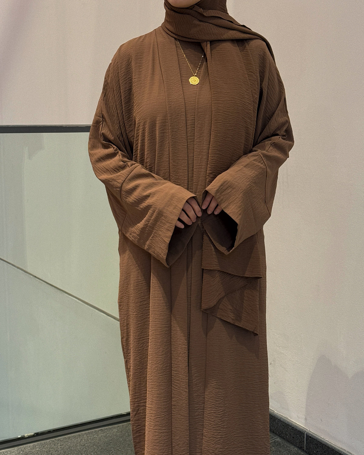 Person wearing a brown long-sleeve dress with a matching hijab against a neutral background