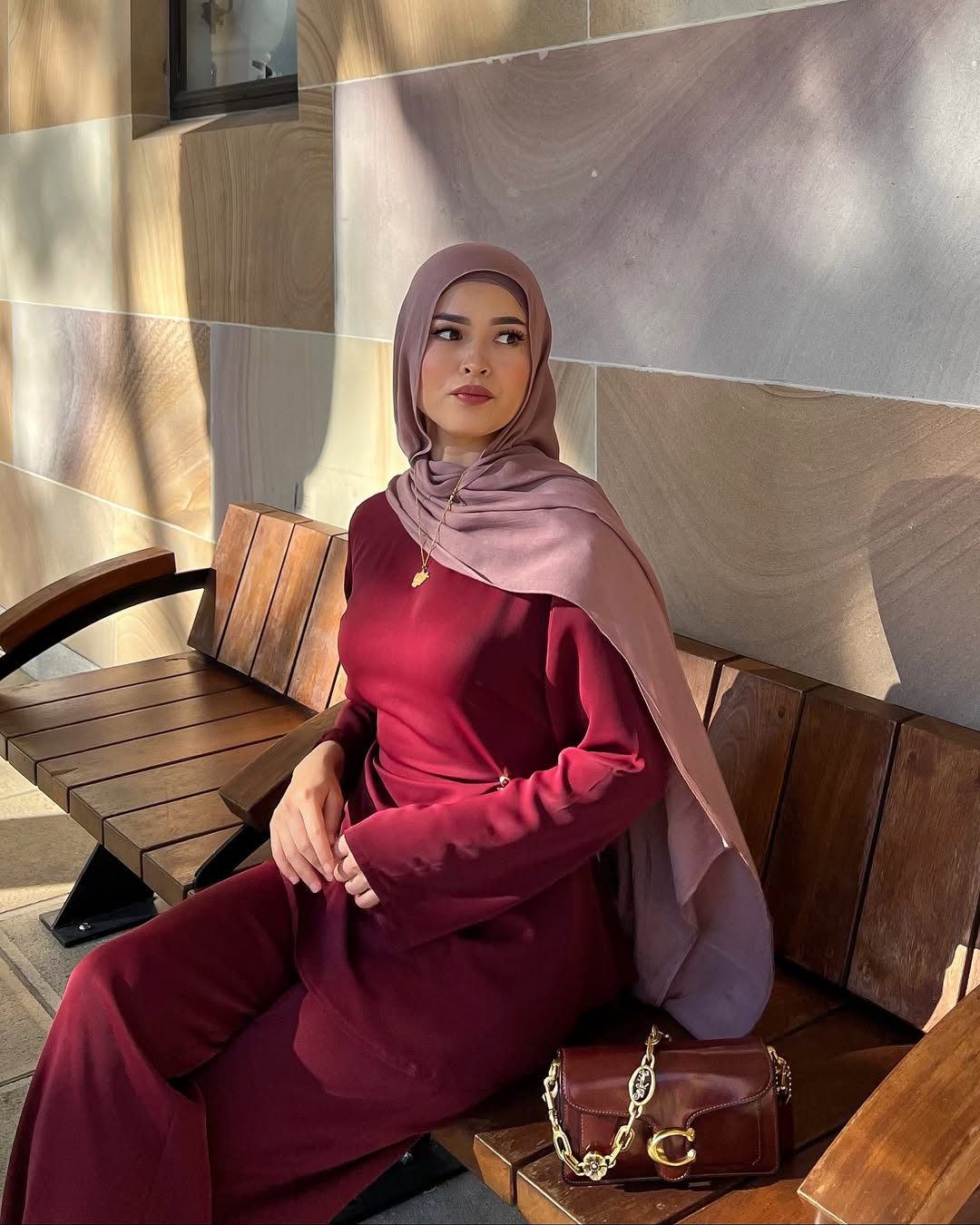 Woman in a red outfit and pink hijab sitting on a wooden bench with a handbag.