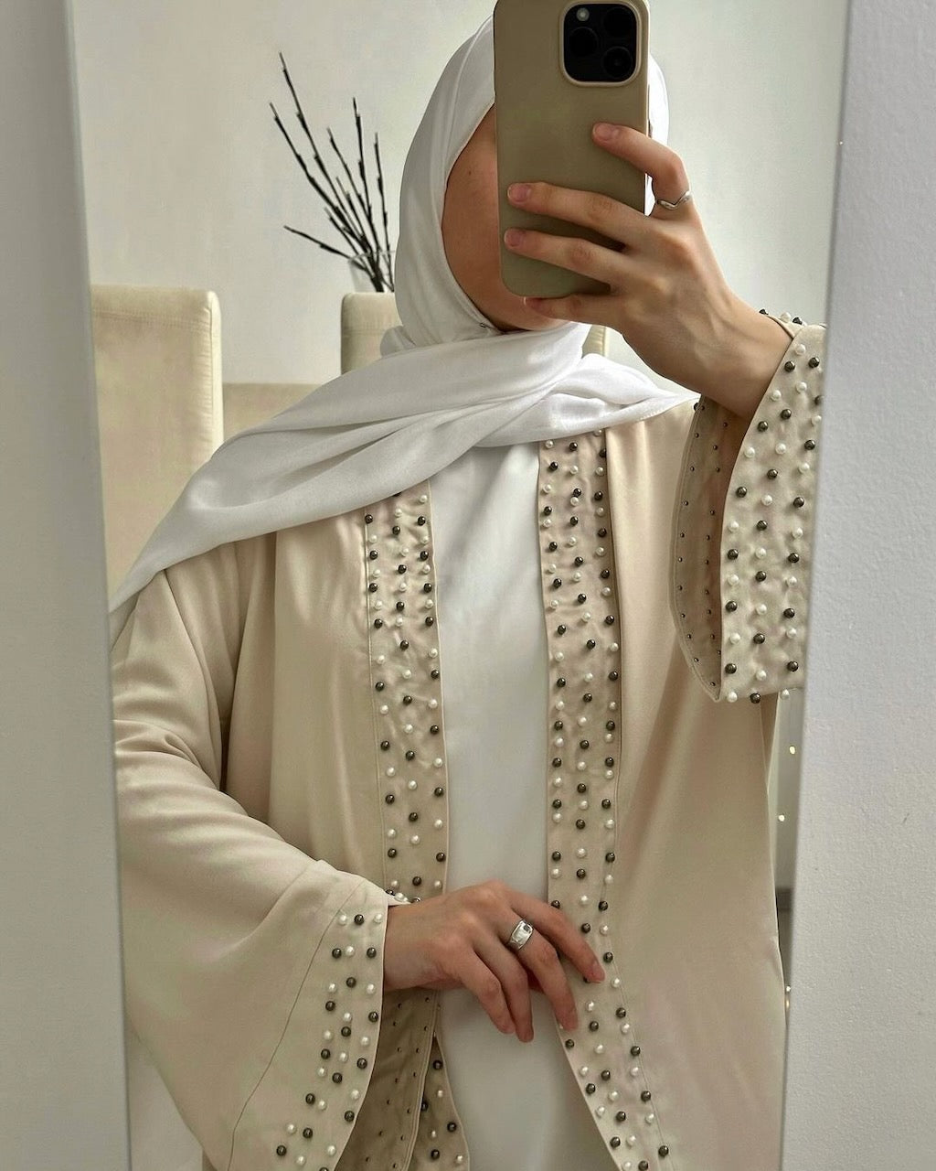 A woman takes a mirror selfie wearing a cream-colored abaya with silver bead embellishments along the front panels and cuffs, paired with a white hijab.