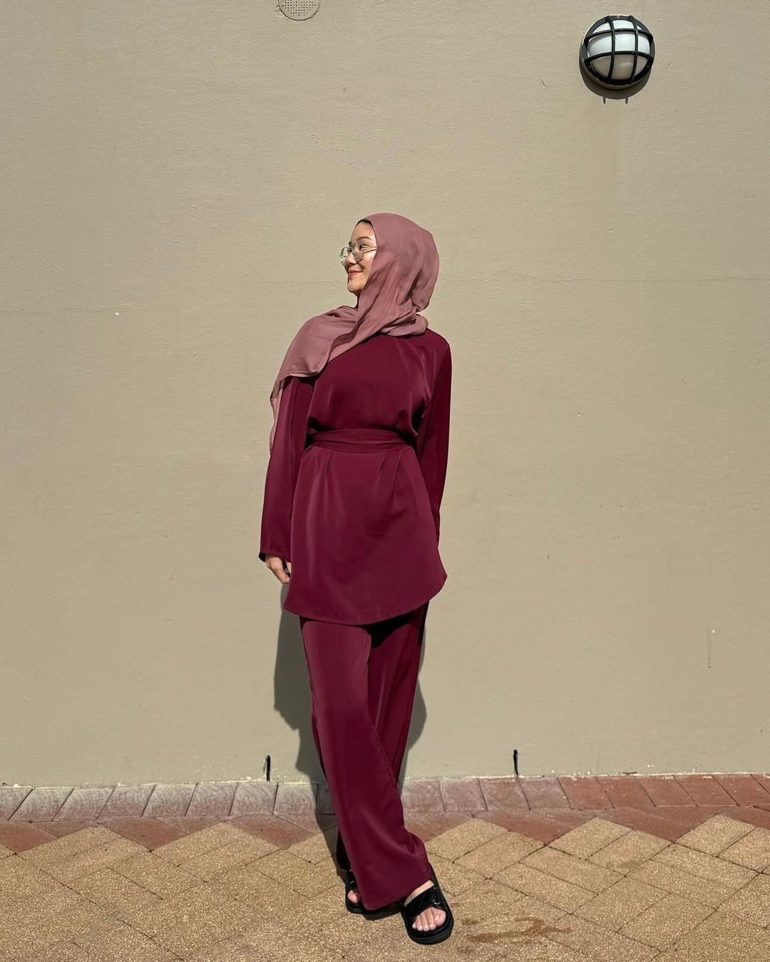Person wearing a maroon outfit with a hijab against a beige wall.