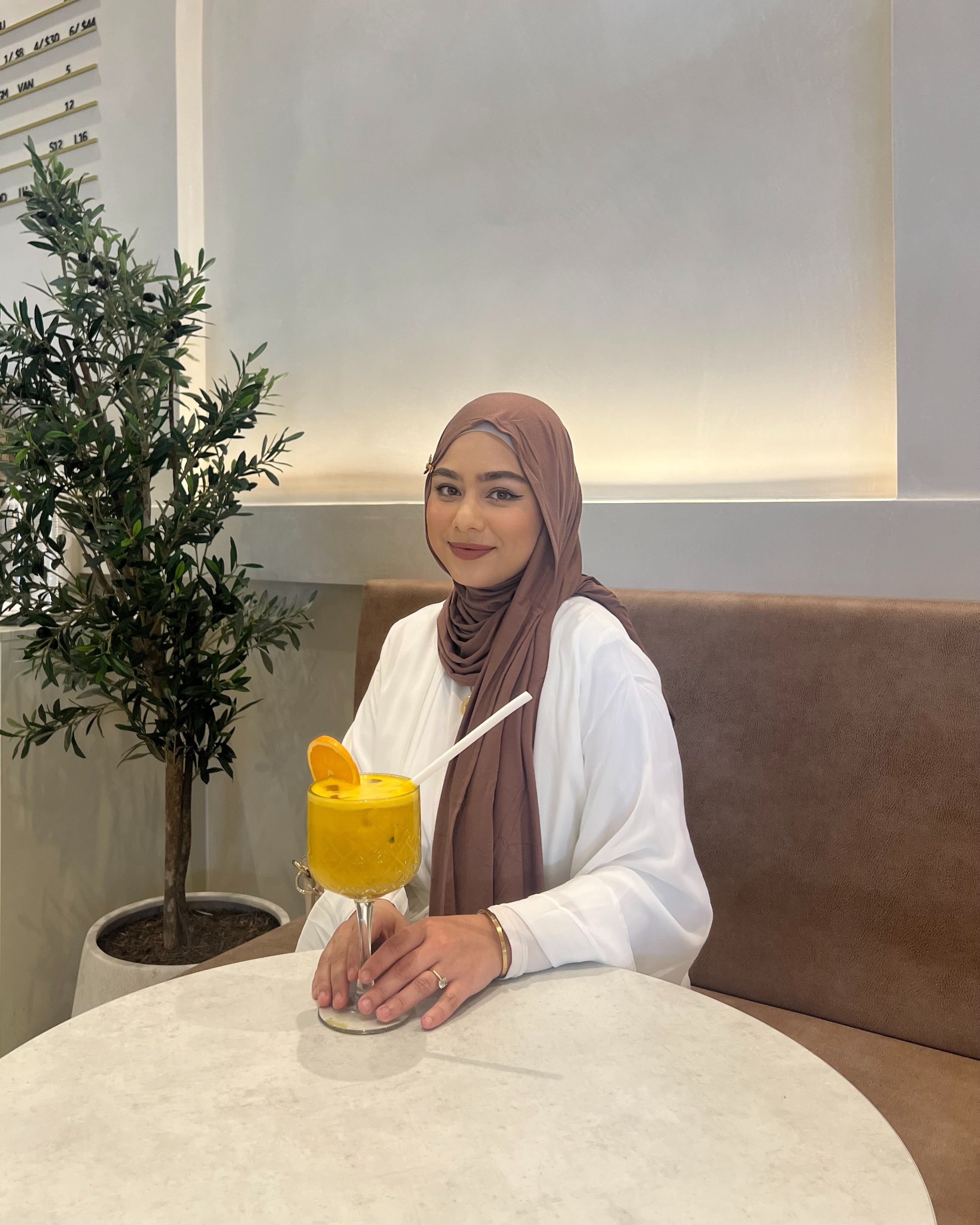 Person wearing a white dress and brown hijab holding a mango orange drink in a room with sofa and table.