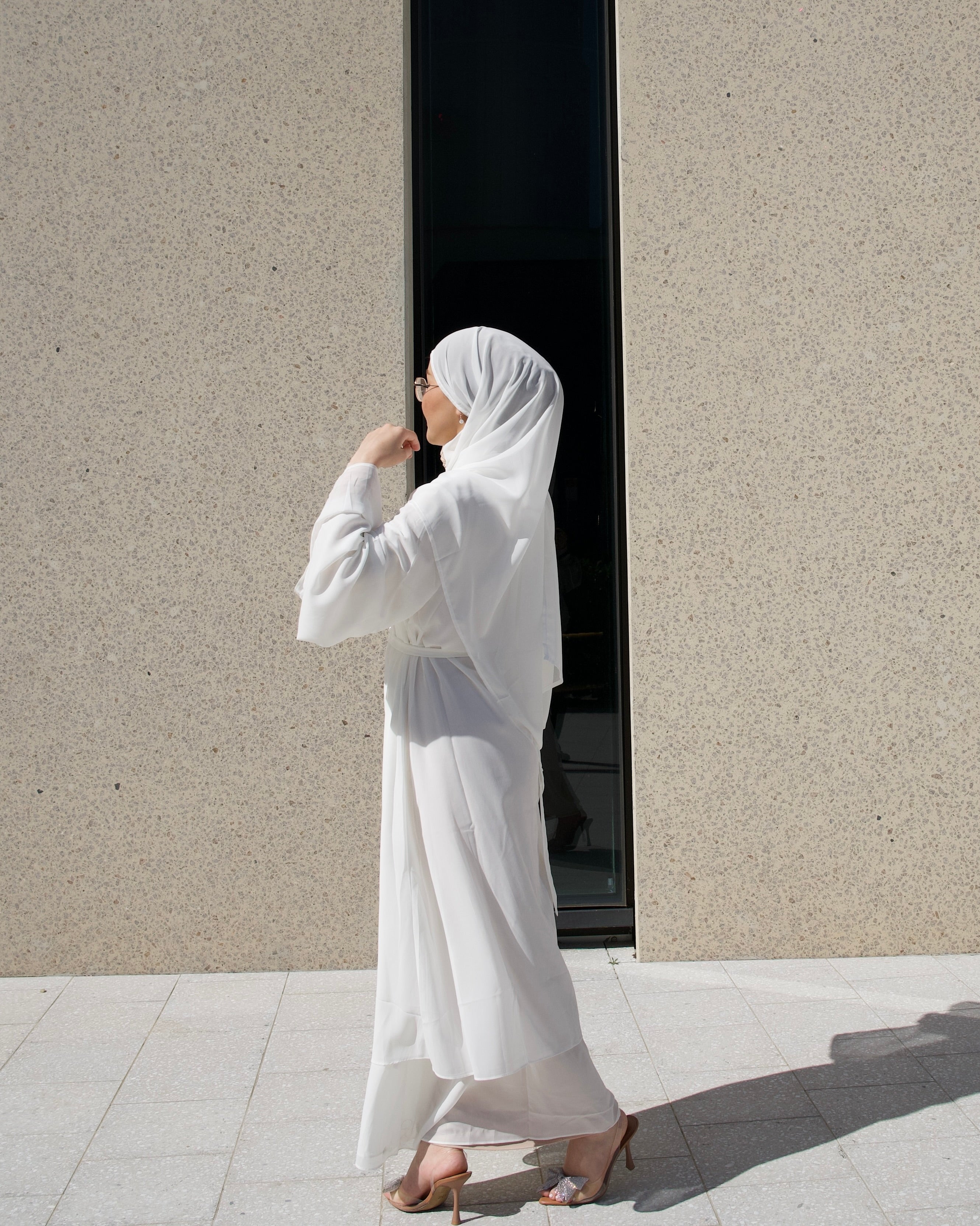 Woman in a white hijab and dress standing against a plain wall.