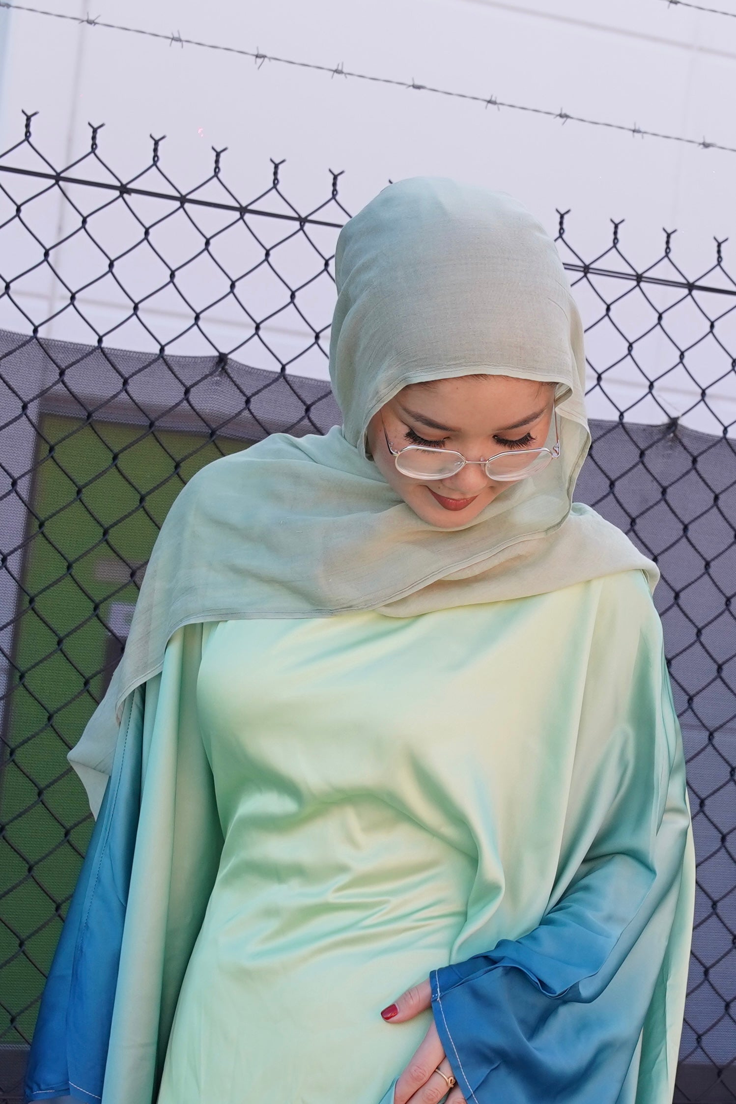 Person wearing a light green hijab and dress with blue sleeves against a chain-link fence background.