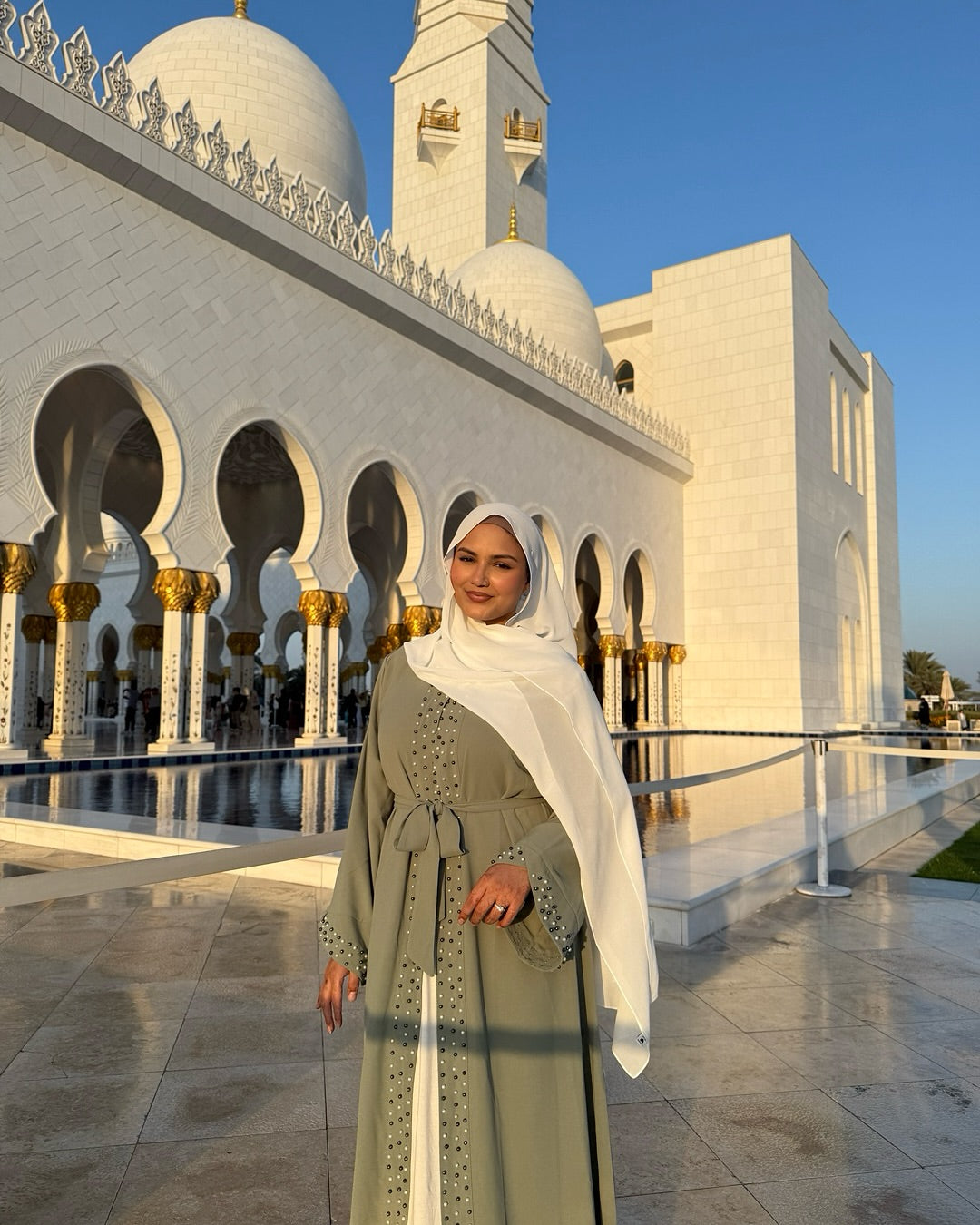 Woman in a green dress and white hijab standing in front of a large, white architectural structure with gold accents.