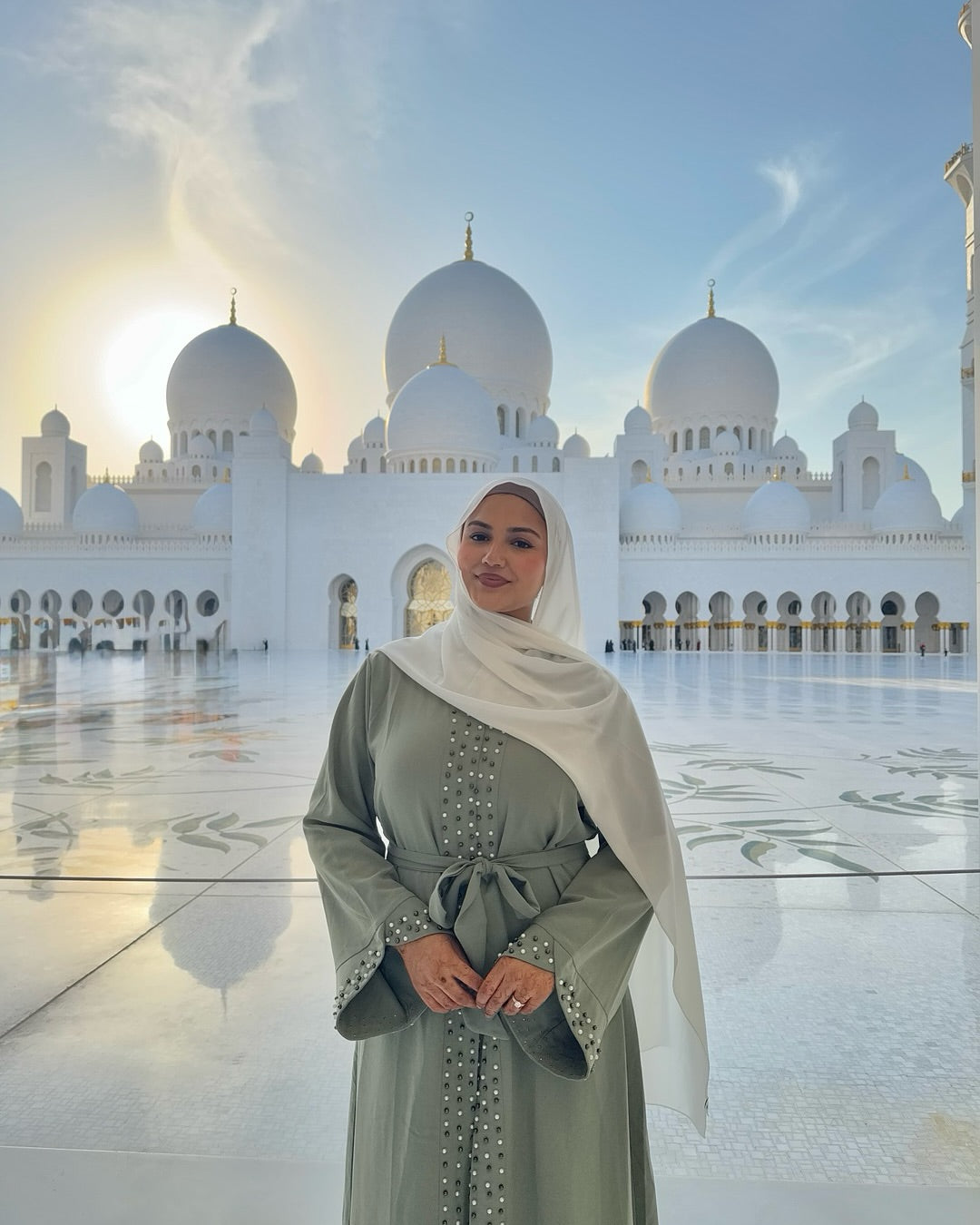 Woman in a light green dress and white hijab standing in front of a mosque with domes.
