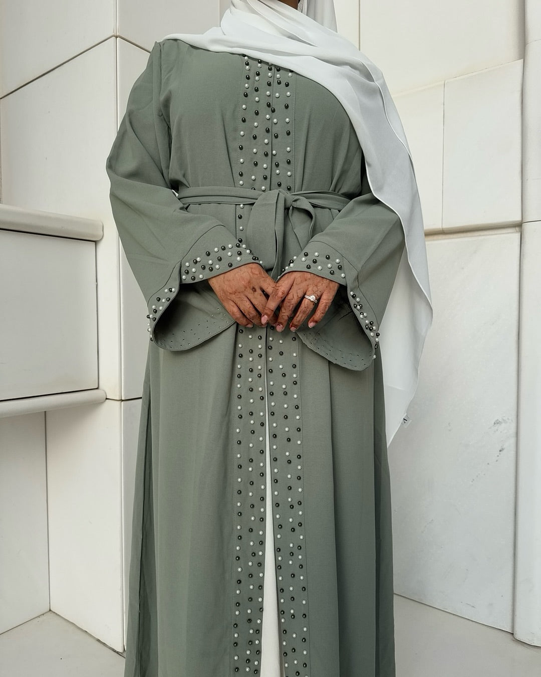 Person wearing a gray abaya with decorative elements in front of a white wall.