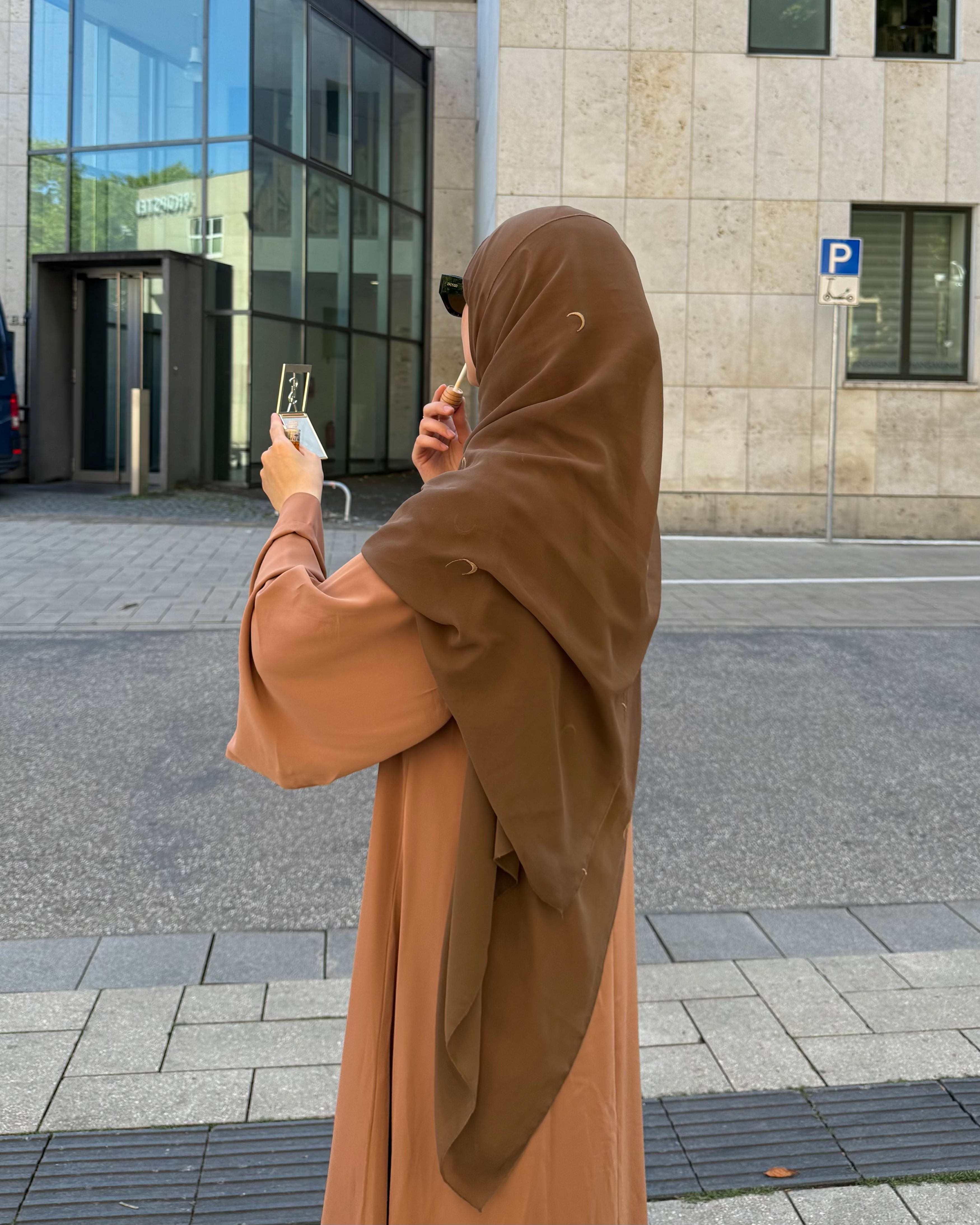 Person in a brown hijab and abaya taking a selfie on a city street.