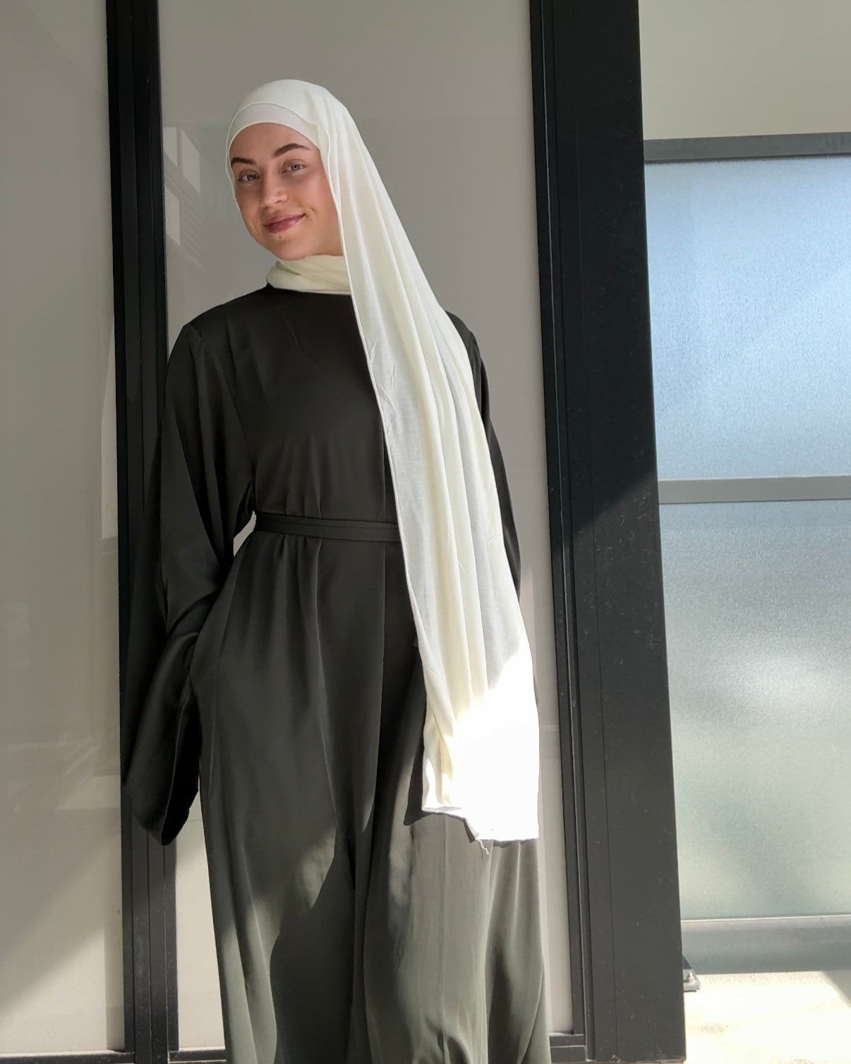 Person wearing a black outfit with a white headscarf standing in front of a glass door.
