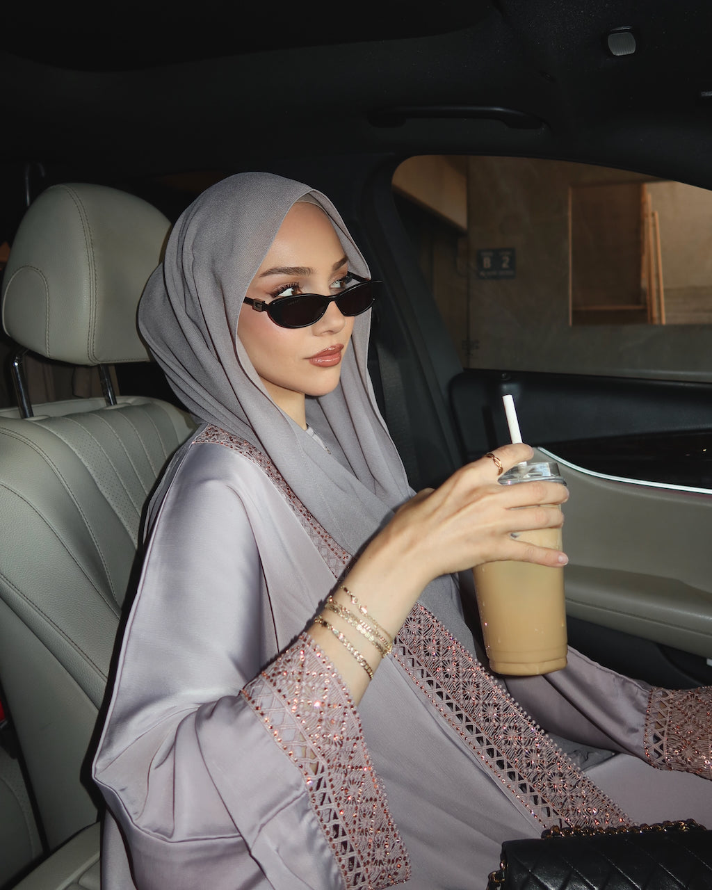 A woman wearing a light gray hijab and a matching embroidered abaya sits in a car, holding an iced coffee. - Fajr Noor