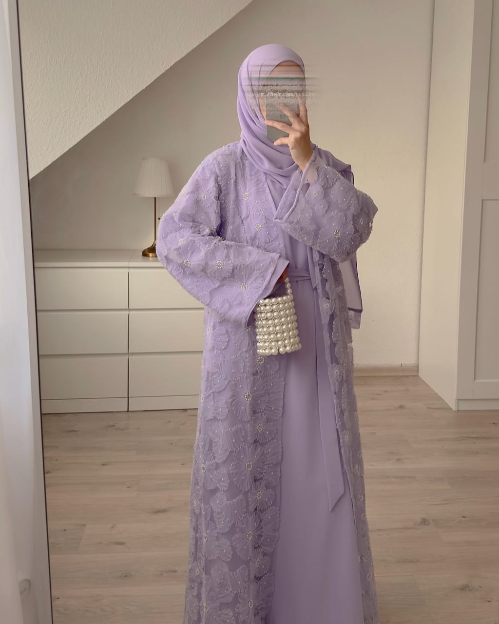 A person wearing a lavender purple modest outfit consisting of a long embroidered abaya, matching hijab, and a pearl-embellished handbag, standing in a mirror selfie in a modern room with light wood floors and white walls.