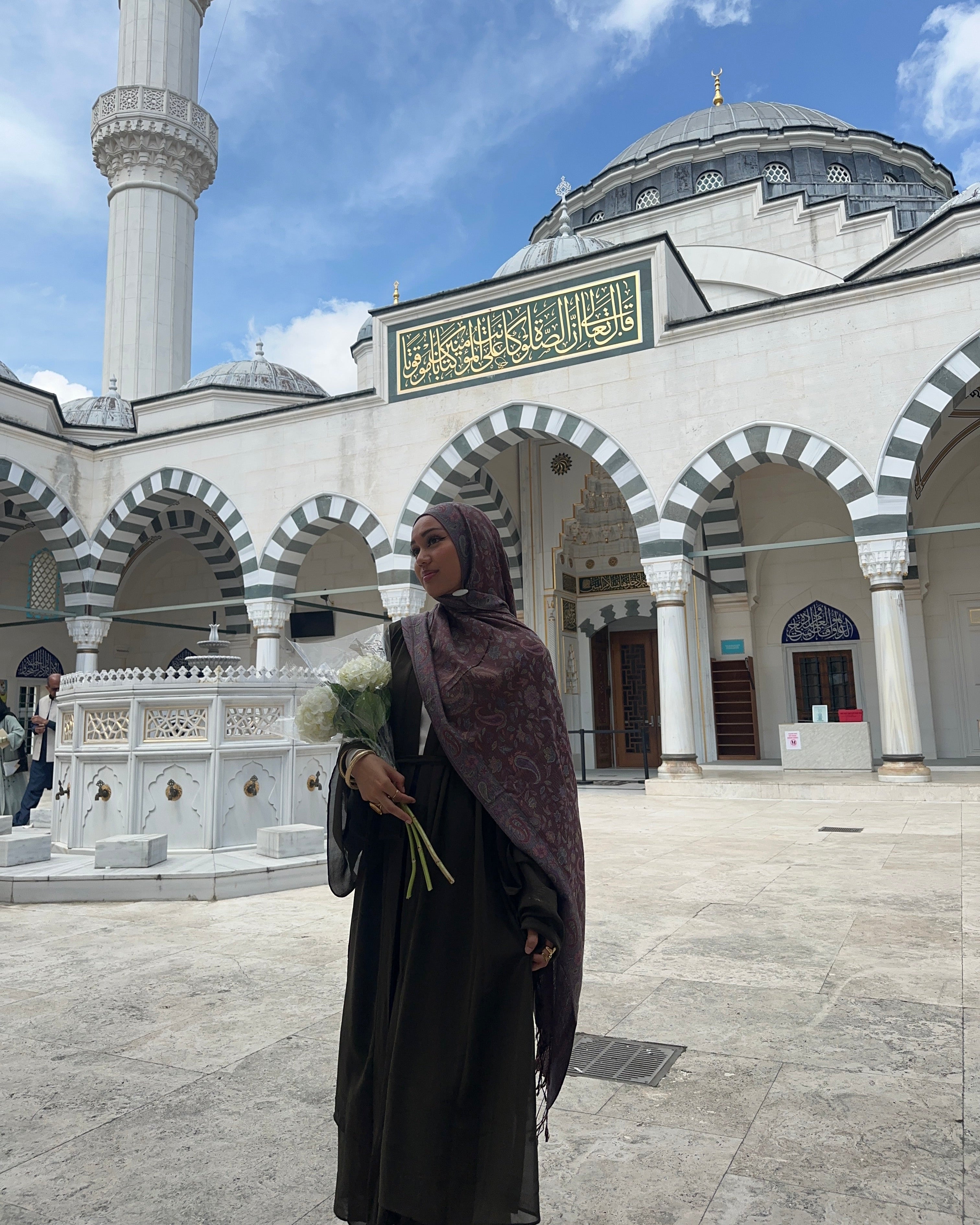 Woman in a hijab standing in front of a mosque with a blue sky.