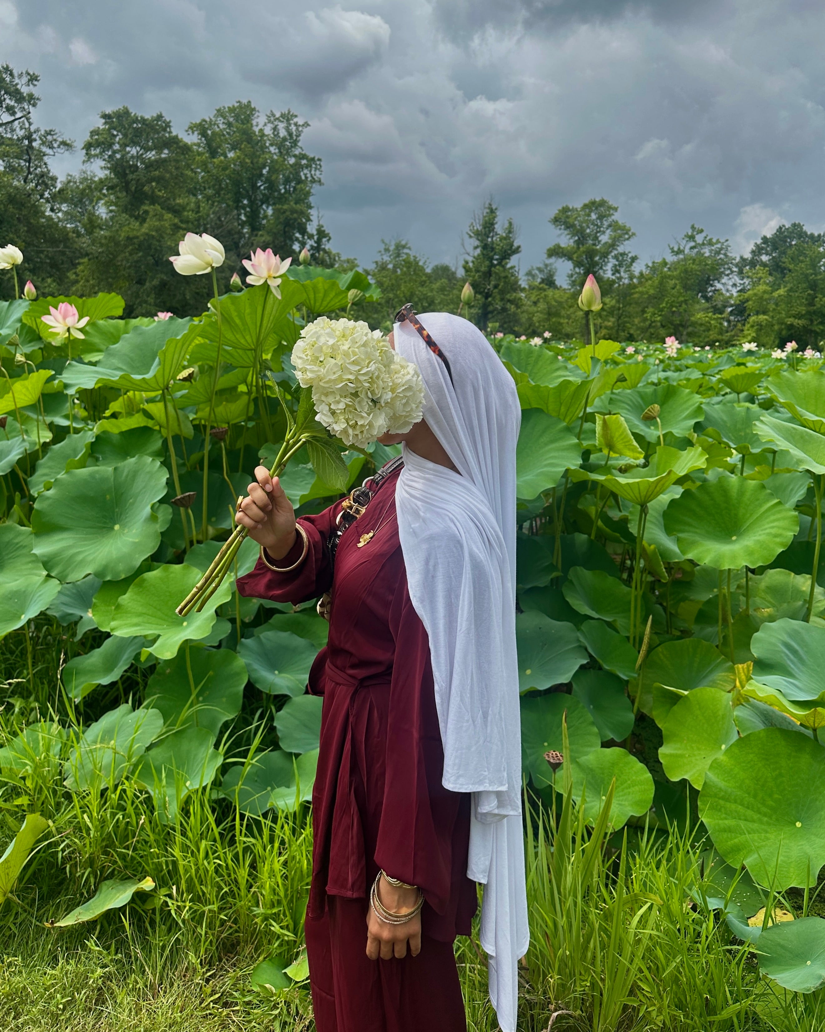 Person holding a bouquet of white flowers amidst a field of lotus plants under a cloudy sky.