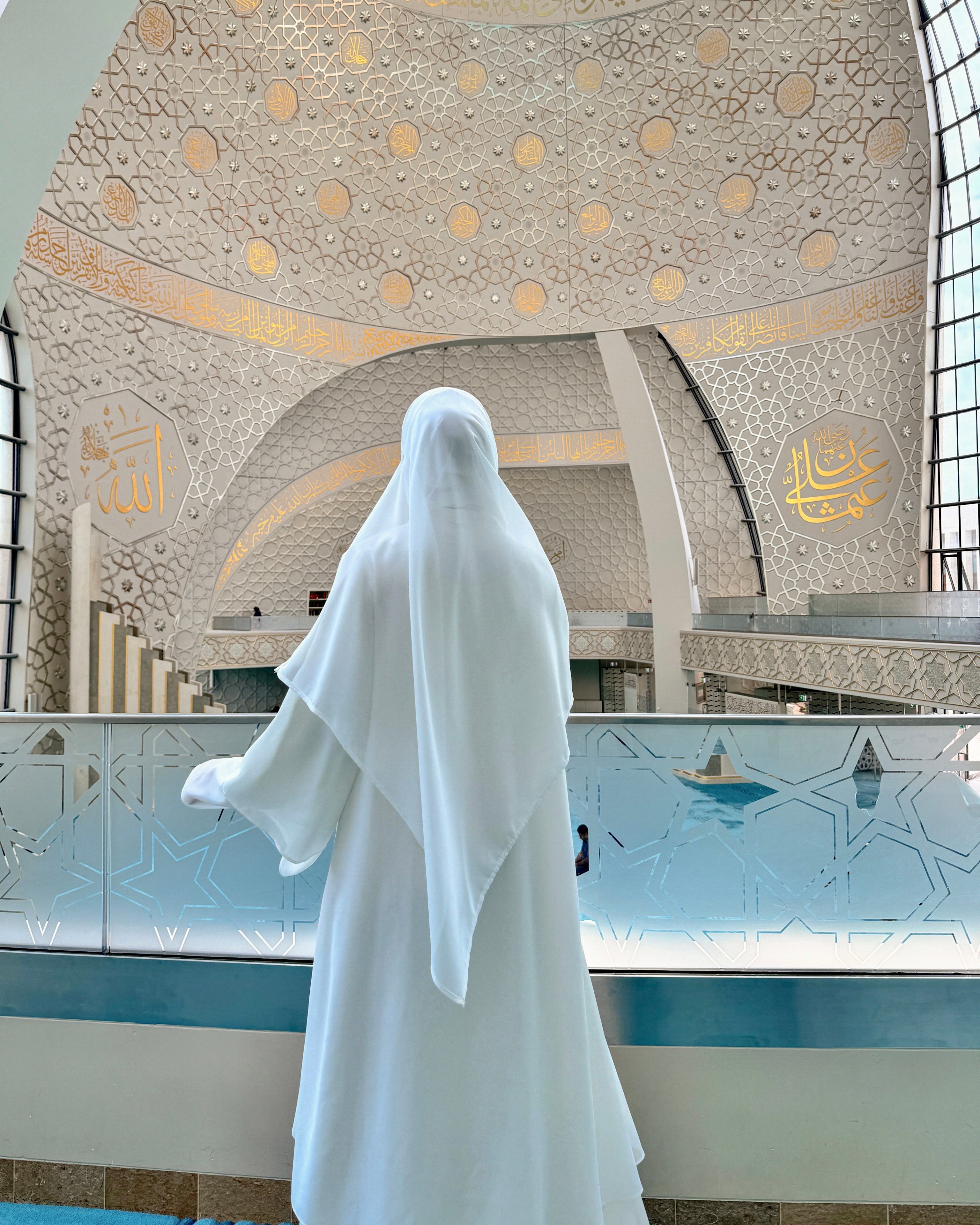 Person in a white dress standing in front of a decorated ceiling inside a mosque building.