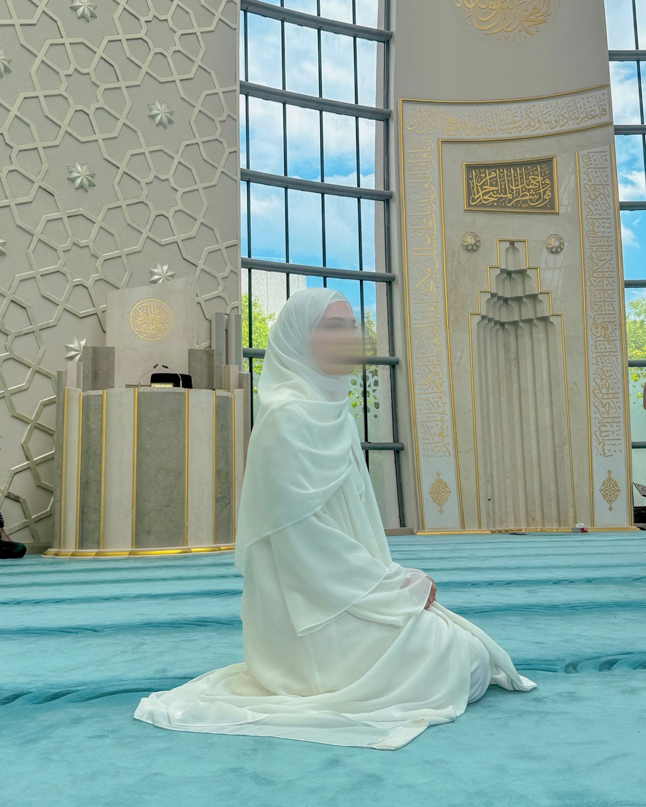 Person in a white outfit kneeling on a blue carpet in a mosque.