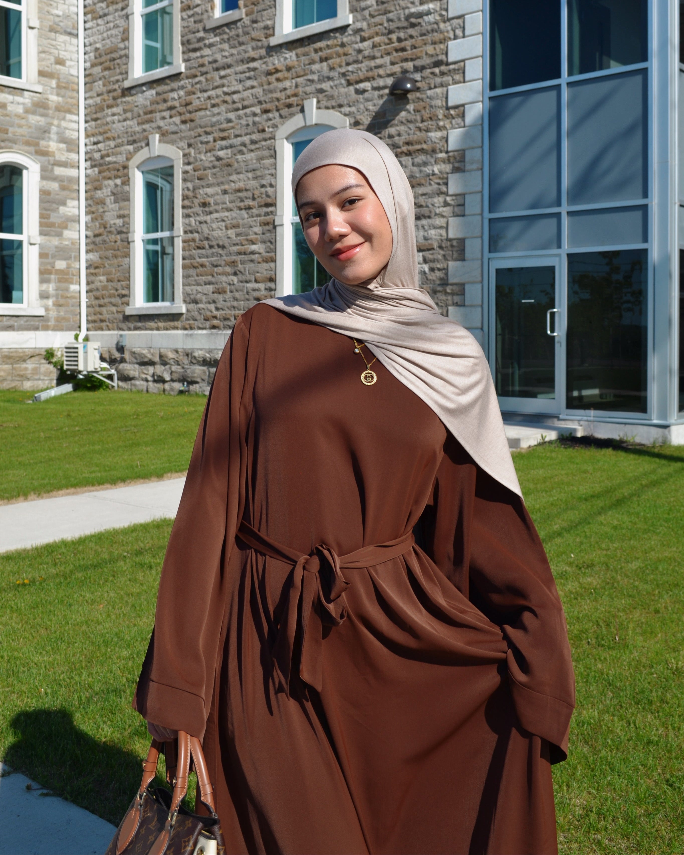 Person wearing a brown outfit and headscarf standing in front of a building with stone and glass walls.