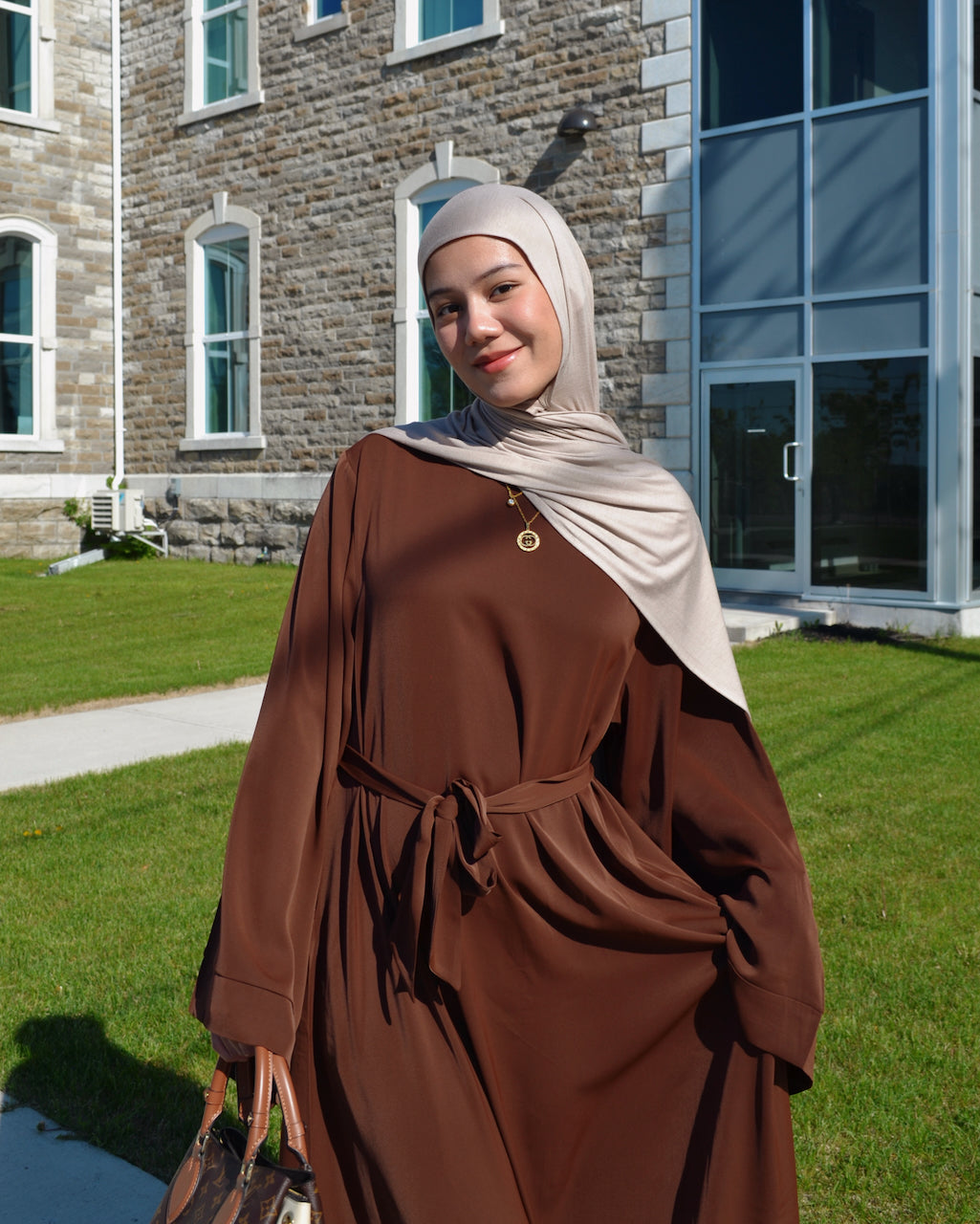 A woman wearing a brown long-sleeve abaya dress with a tie waist and a light beige hijab, standing outdoors in front of a stone building and green lawn.