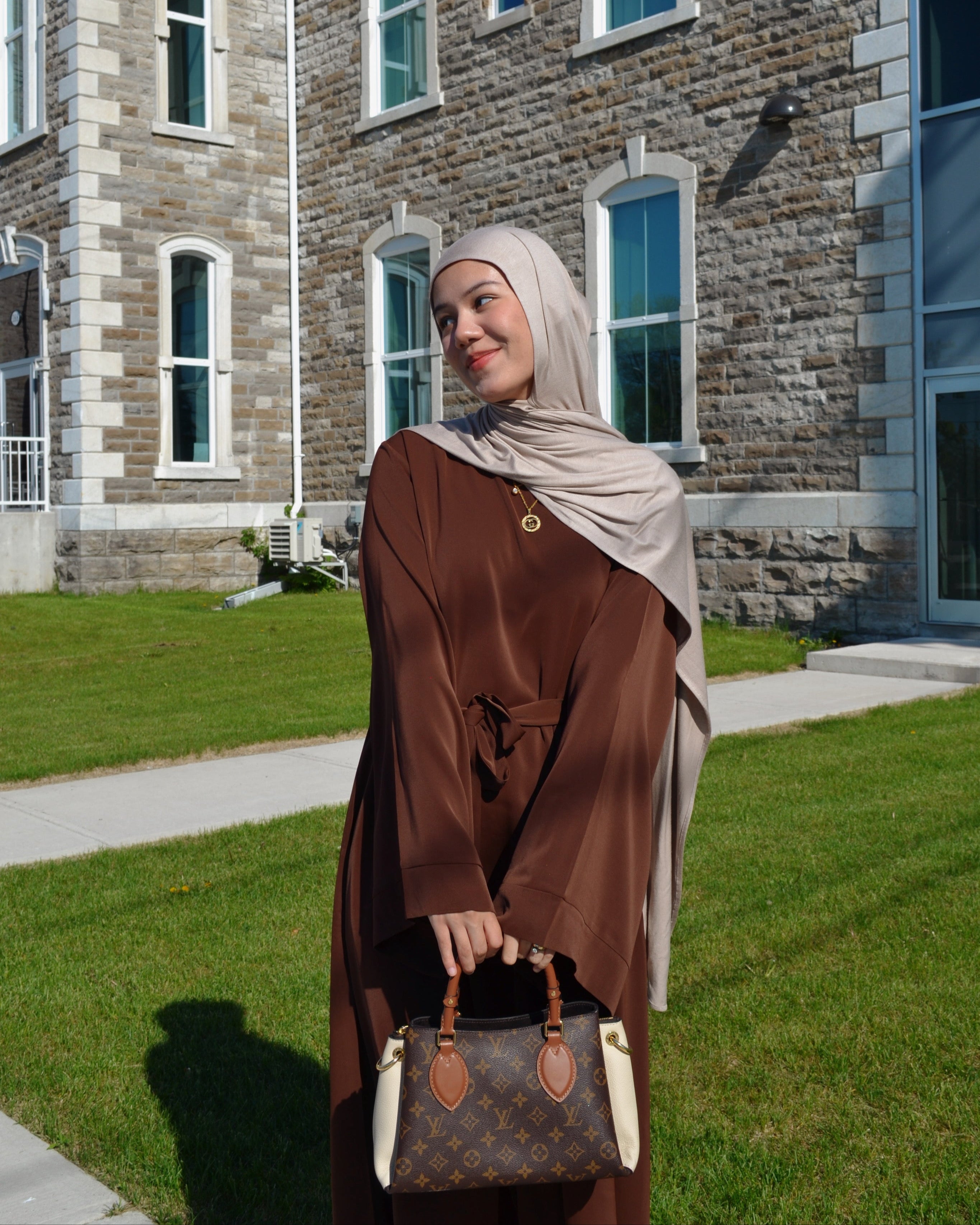 Woman in a brown outfit with a headscarf standing in front of a stone building.