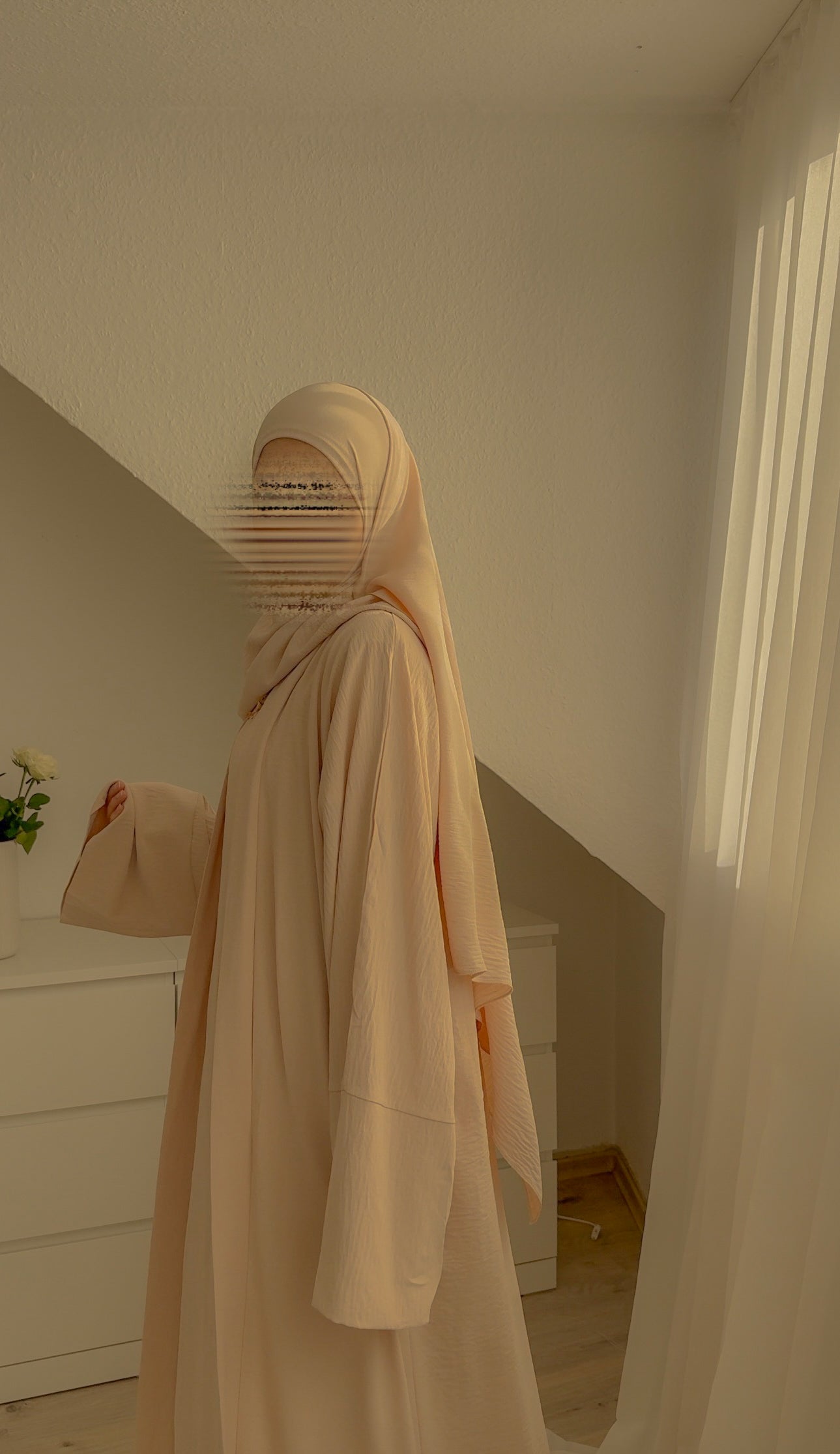Person wearing a beige hijab and dress in a minimalistic room.