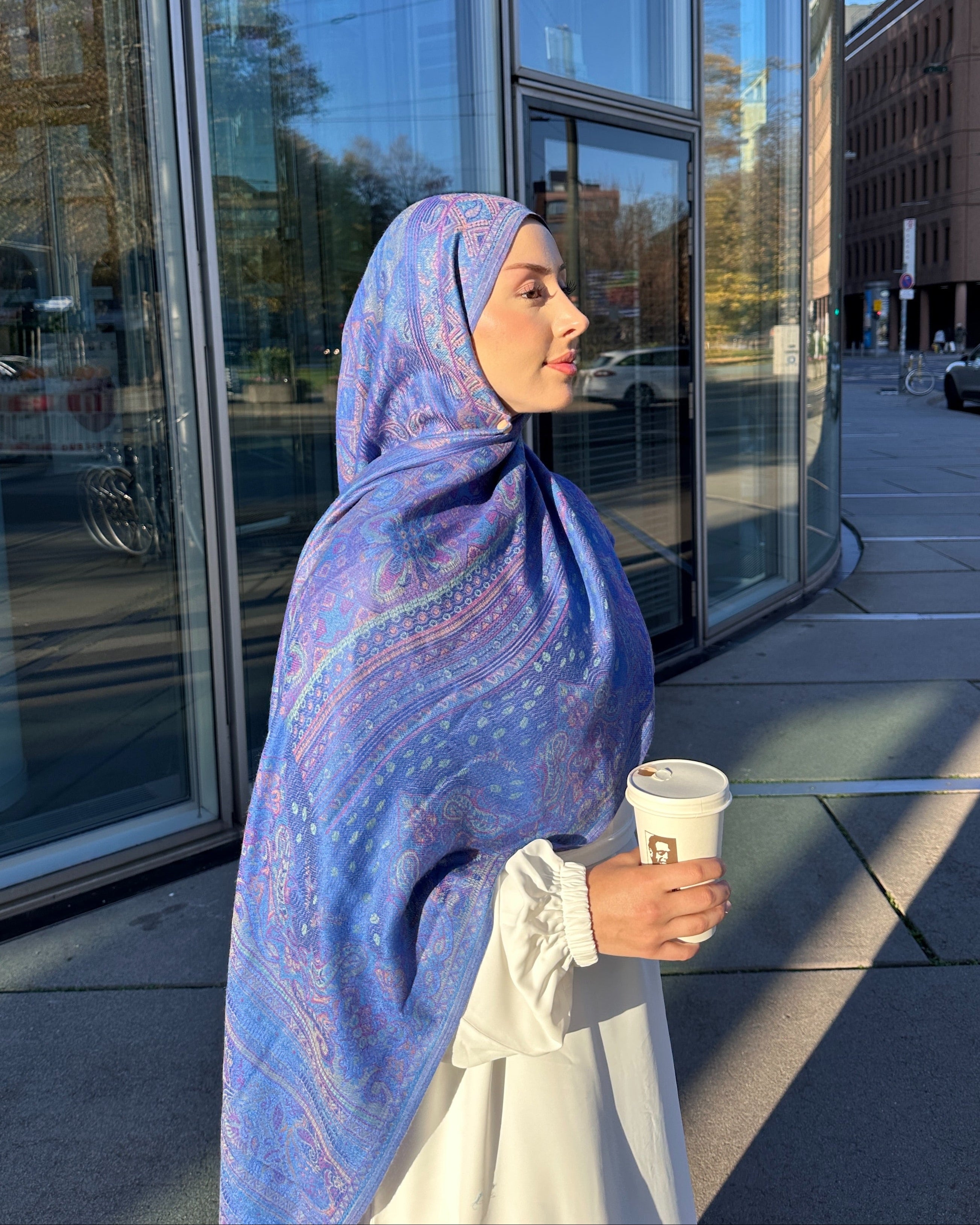 Woman wearing a blue patterned hijab holding a coffee cup outside a building.