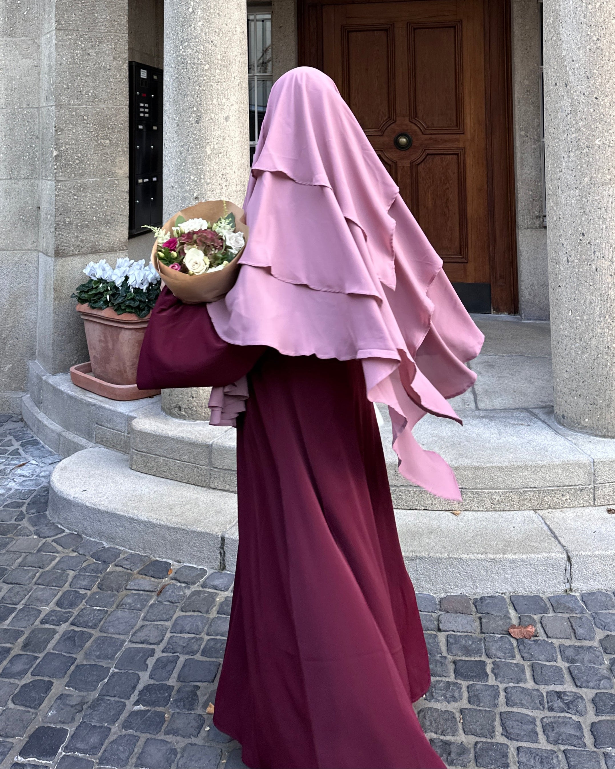 Person wearing a pink hijab and maroon dress walking past a building entrance.