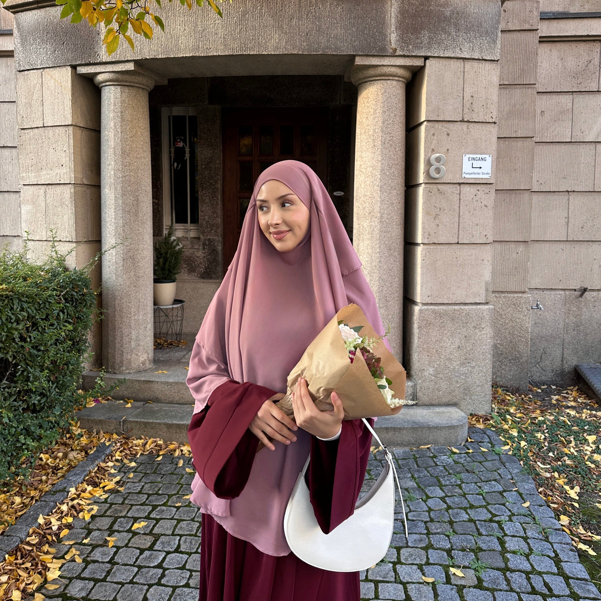 Woman in a pink hijab holding flowers and a white bag outside a building with stone columns.