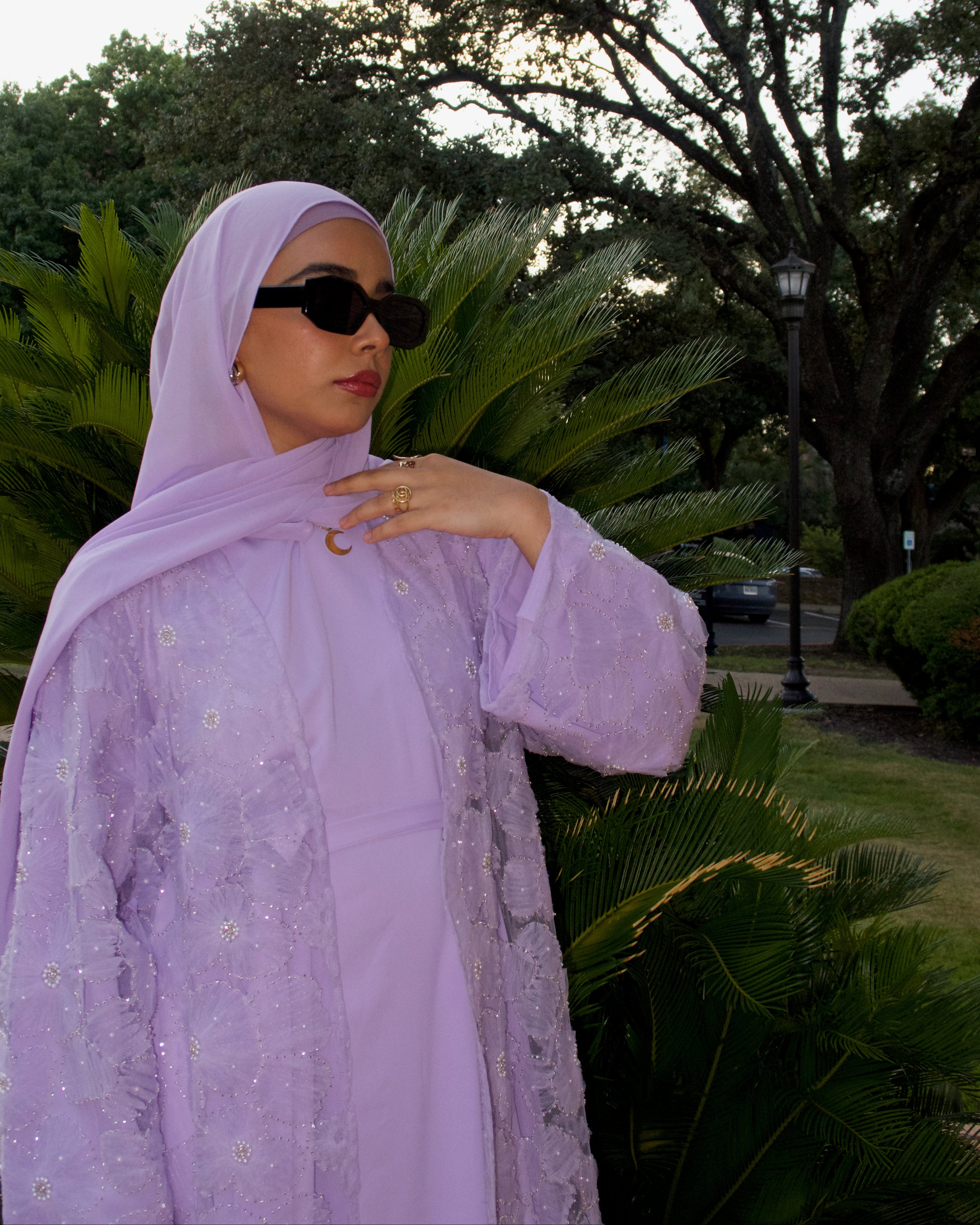 Woman wearing a light purple outfit with a headscarf in an outdoor setting with greenery.