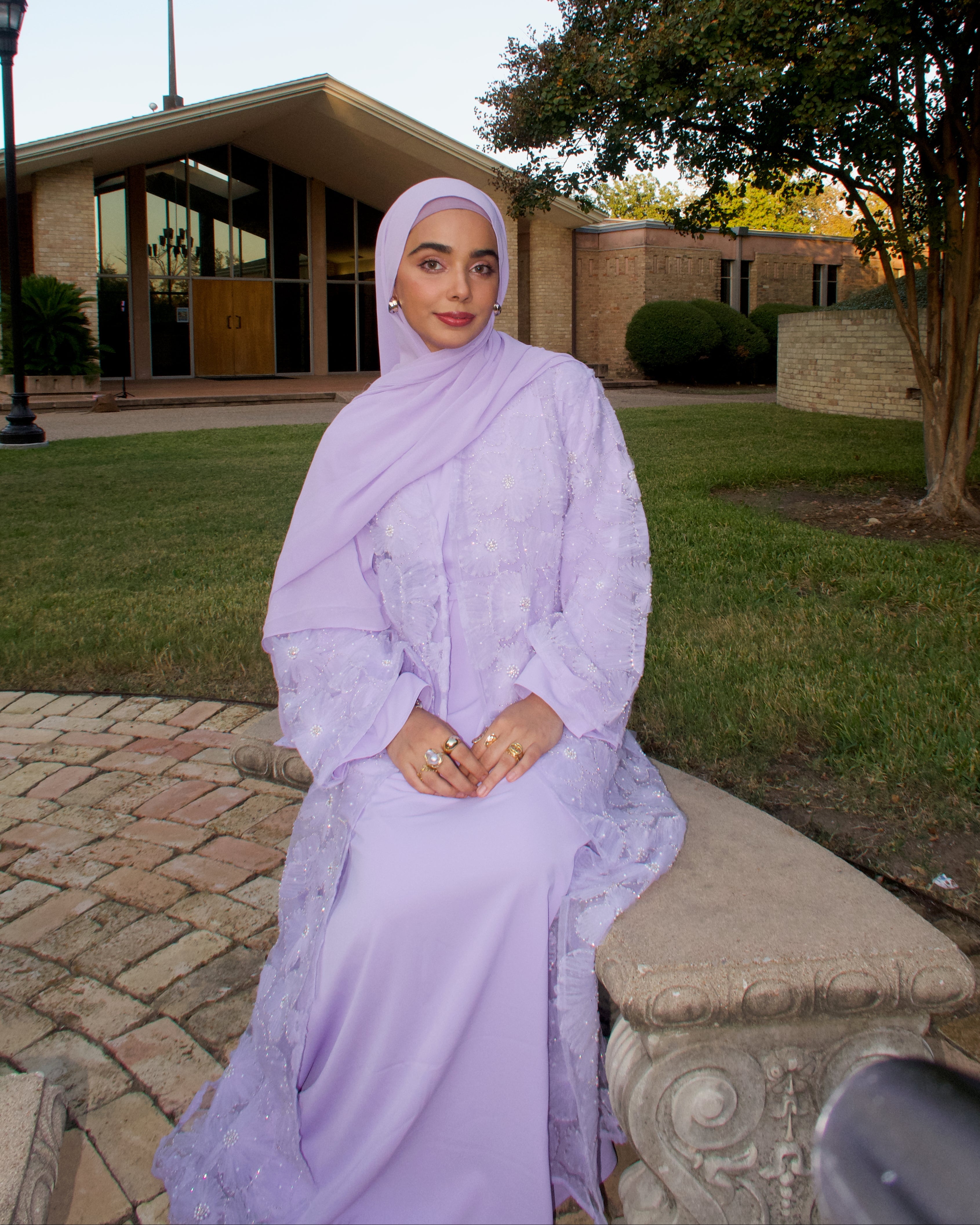Woman in a light purple dress sitting on a bench outdoors with a building and trees in the background.