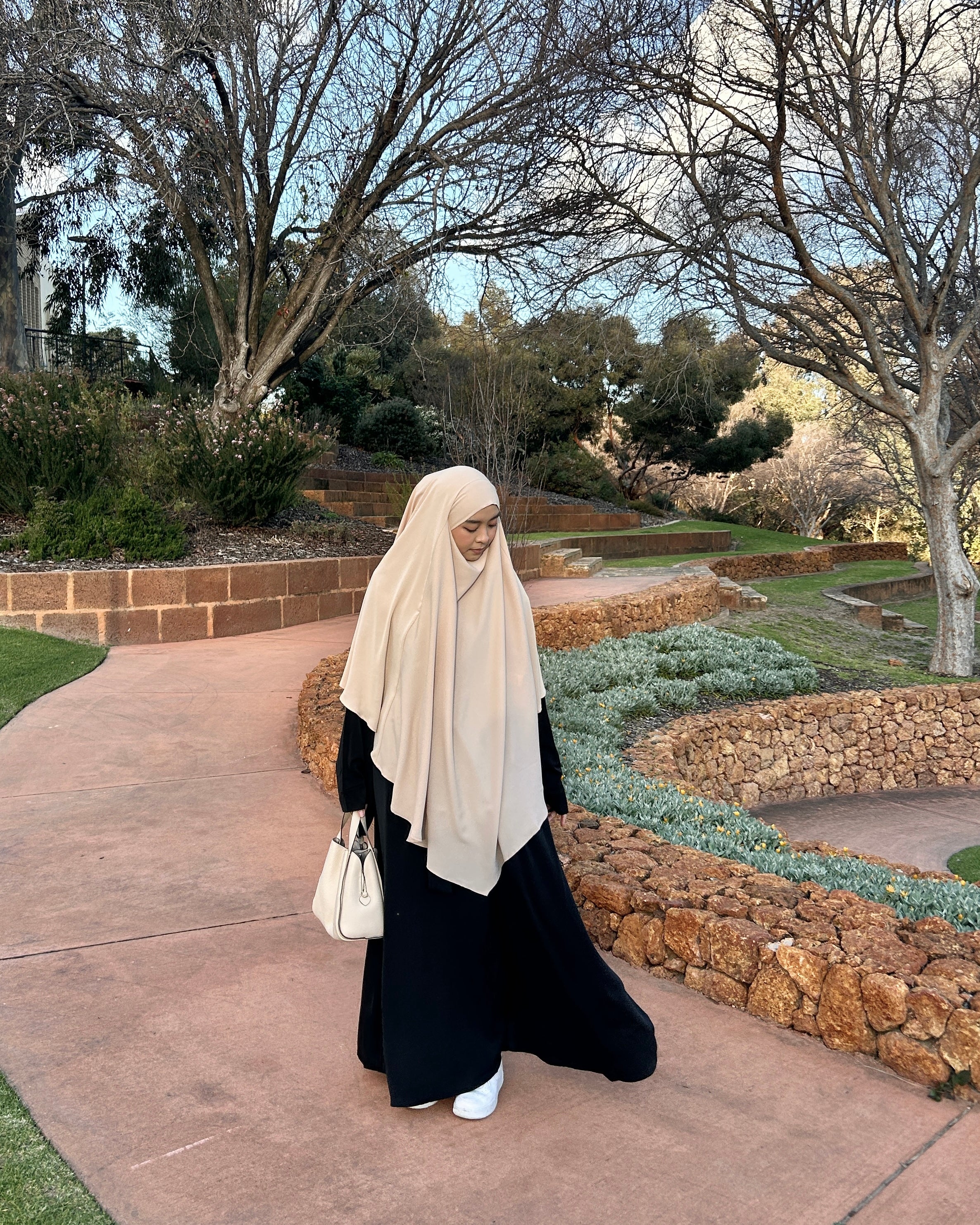 Person wearing a beige hijab and black outfit walking on a path in a garden.