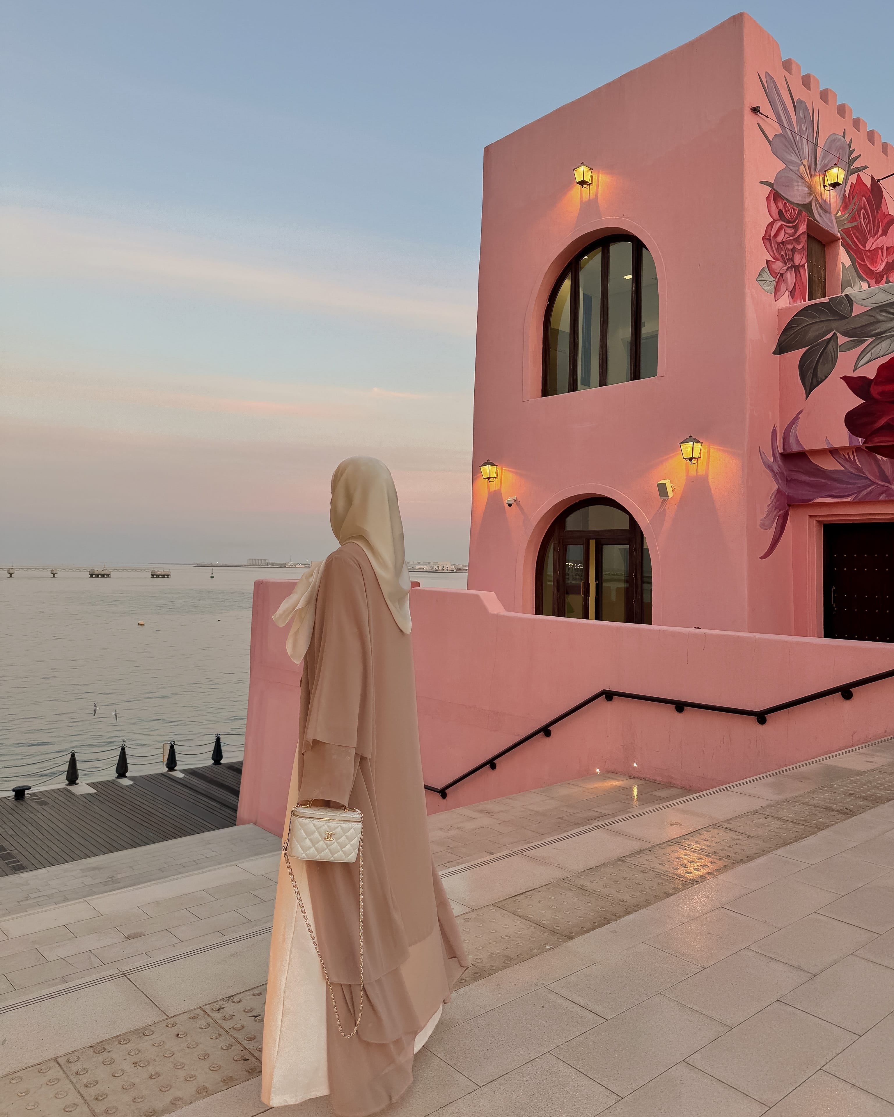 A woman in a beige abaya and hijab stands on a seaside walkway beside a pink building with floral wall art, looking out toward the water at sunset.