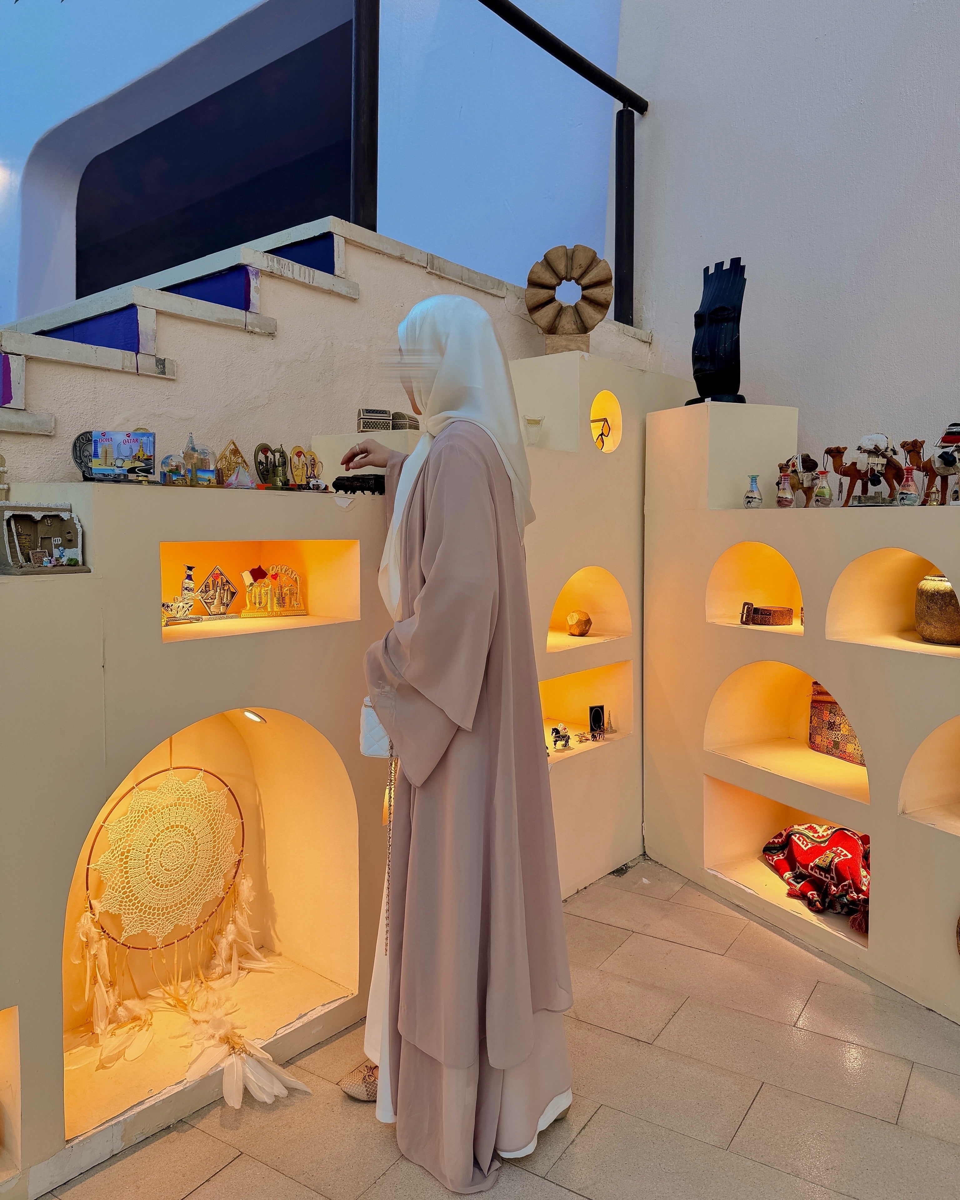 A woman wearing a long beige abaya and white headscarf stands in a boutique interior with cream-colored shelving and warm lighting, surrounded by decorative items.