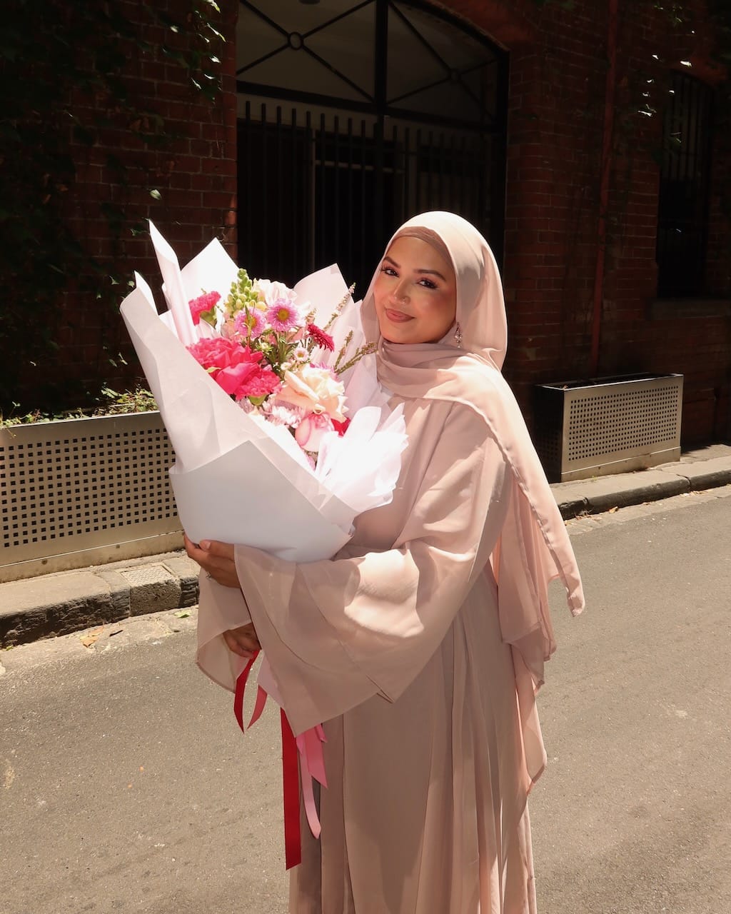 A woman wearing a light pink hijab and matching long-sleeve dress stands outdoors holding a large bouquet of pink and white flowers wrapped in white paper.