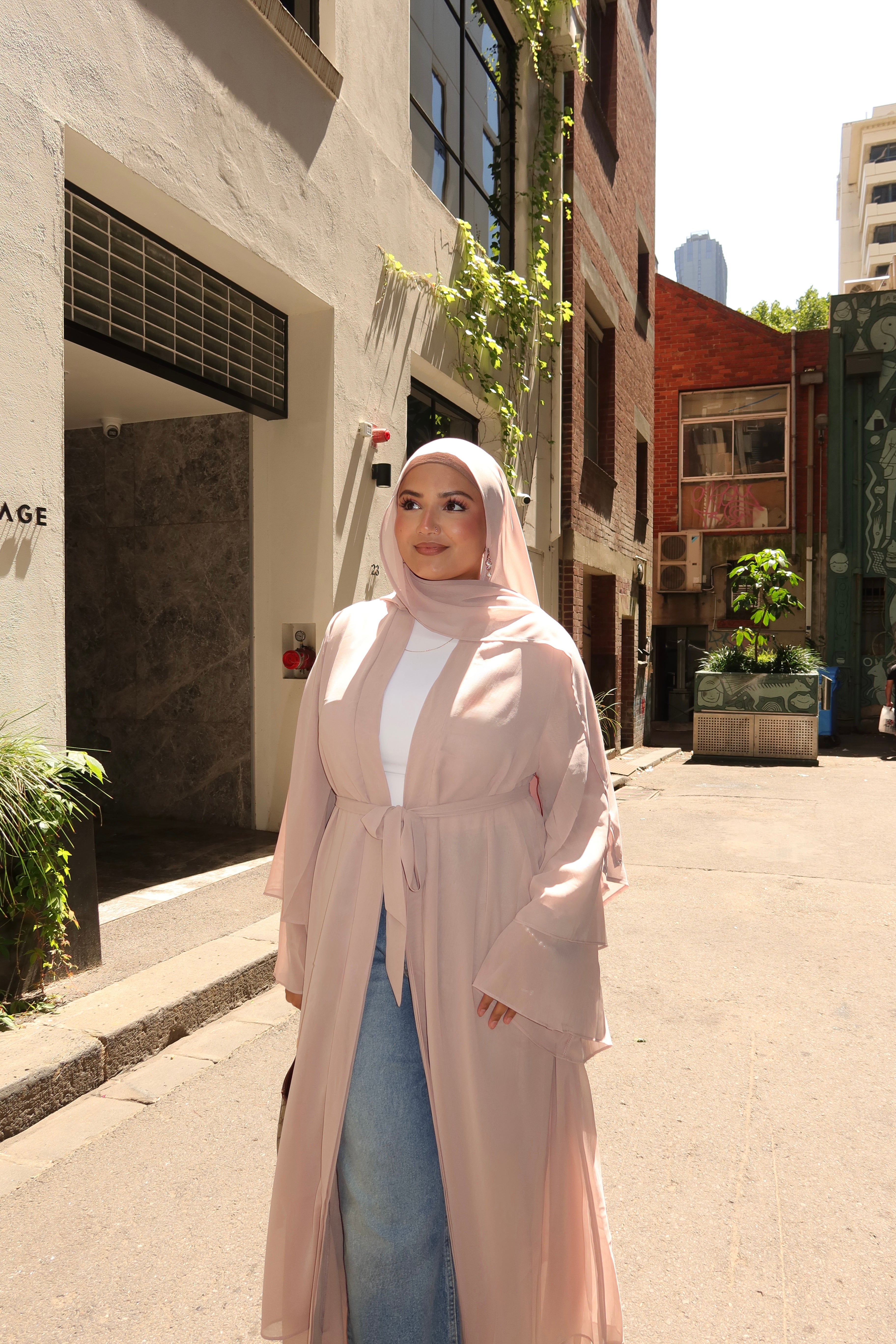 A woman wearing a light pink hijab and a matching long-sleeve, tie-front abaya coat over a white top and blue jeans, standing on a sunlit city street.