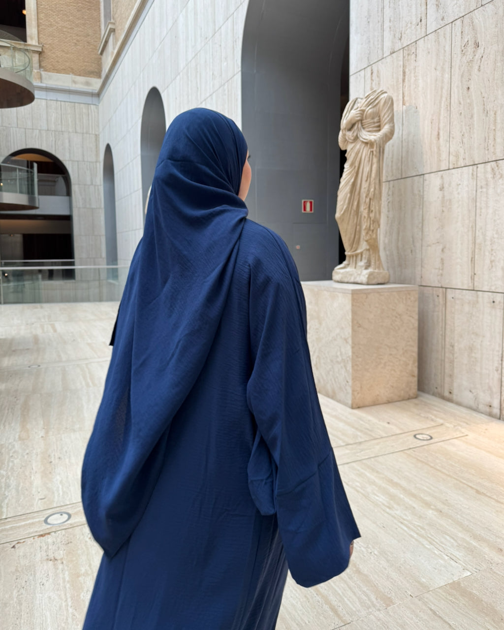 A woman wearing a navy blue abaya and matching hijab stands in a modern stone courtyard, viewed from behind as she looks toward a classical statue.