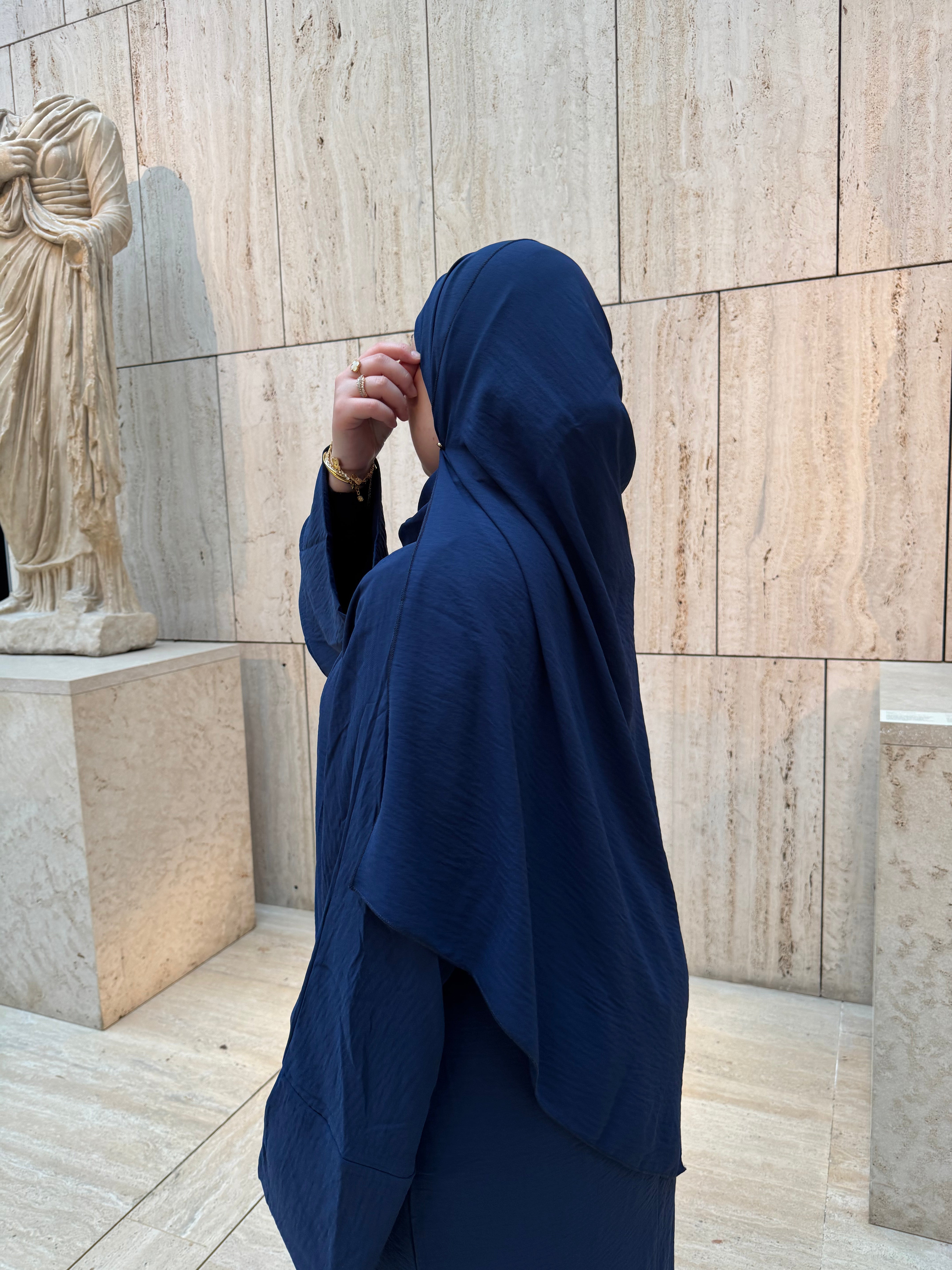 A person wearing a deep navy blue hijab and matching long-sleeve garment stands in profile in front of a beige stone wall, with a classical statue visible on the left.