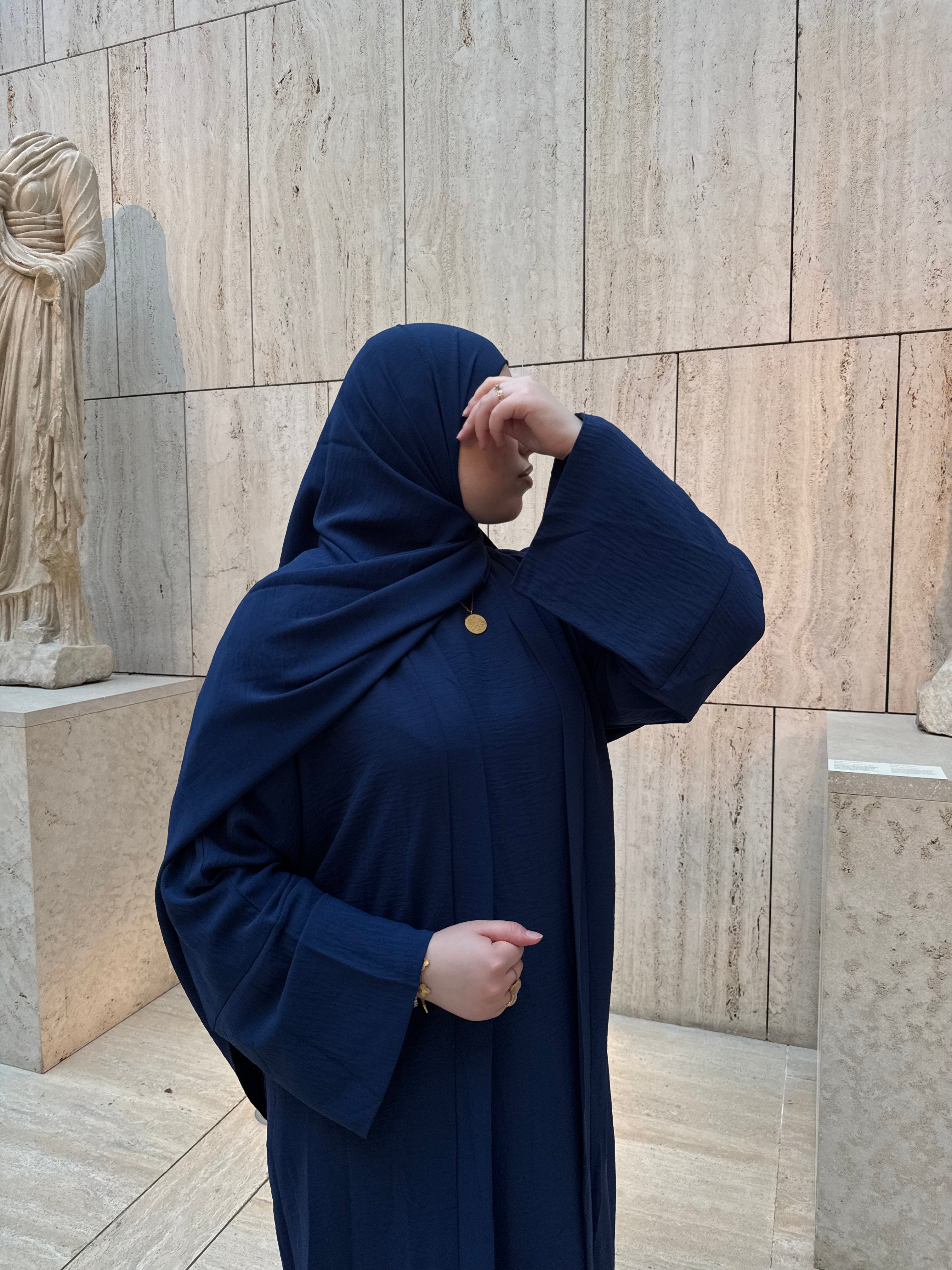 A woman wearing a deep navy blue modest outfit with a matching headscarf and a small gold button detail, standing in front of a beige stone wall.