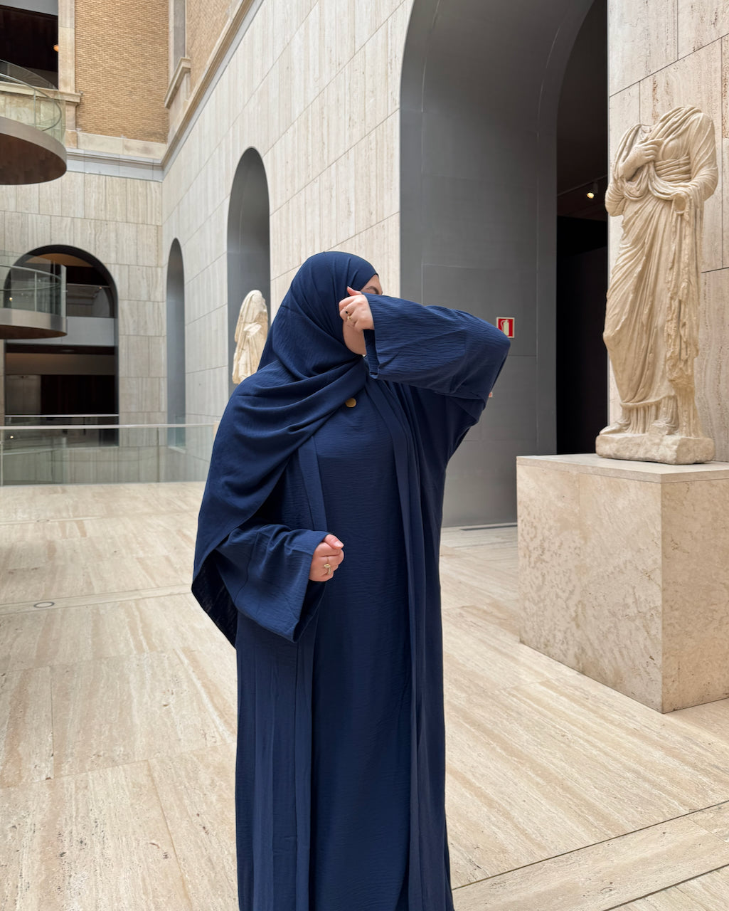 A woman wearing a navy blue modest outfit with a full-length abaya and matching hijab stands in a grand stone courtyard with arches and classical statues.