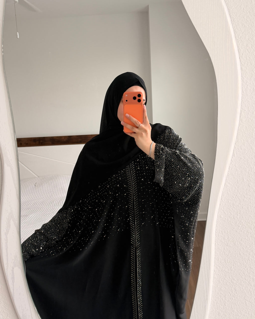 A woman takes a mirror selfie wearing a black abaya with sparkling silver embellishments and a matching black hijab. She is holding an orange smartphone in front of her face.