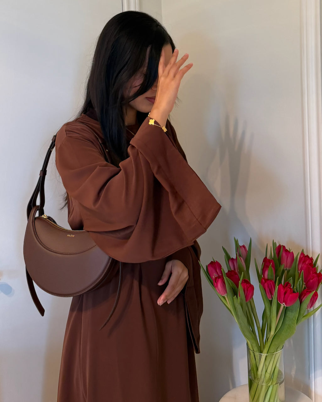 A woman wearing a brown long-sleeve dress and carrying a brown crescent shoulder bag, standing indoors next to a vase of pink tulips.