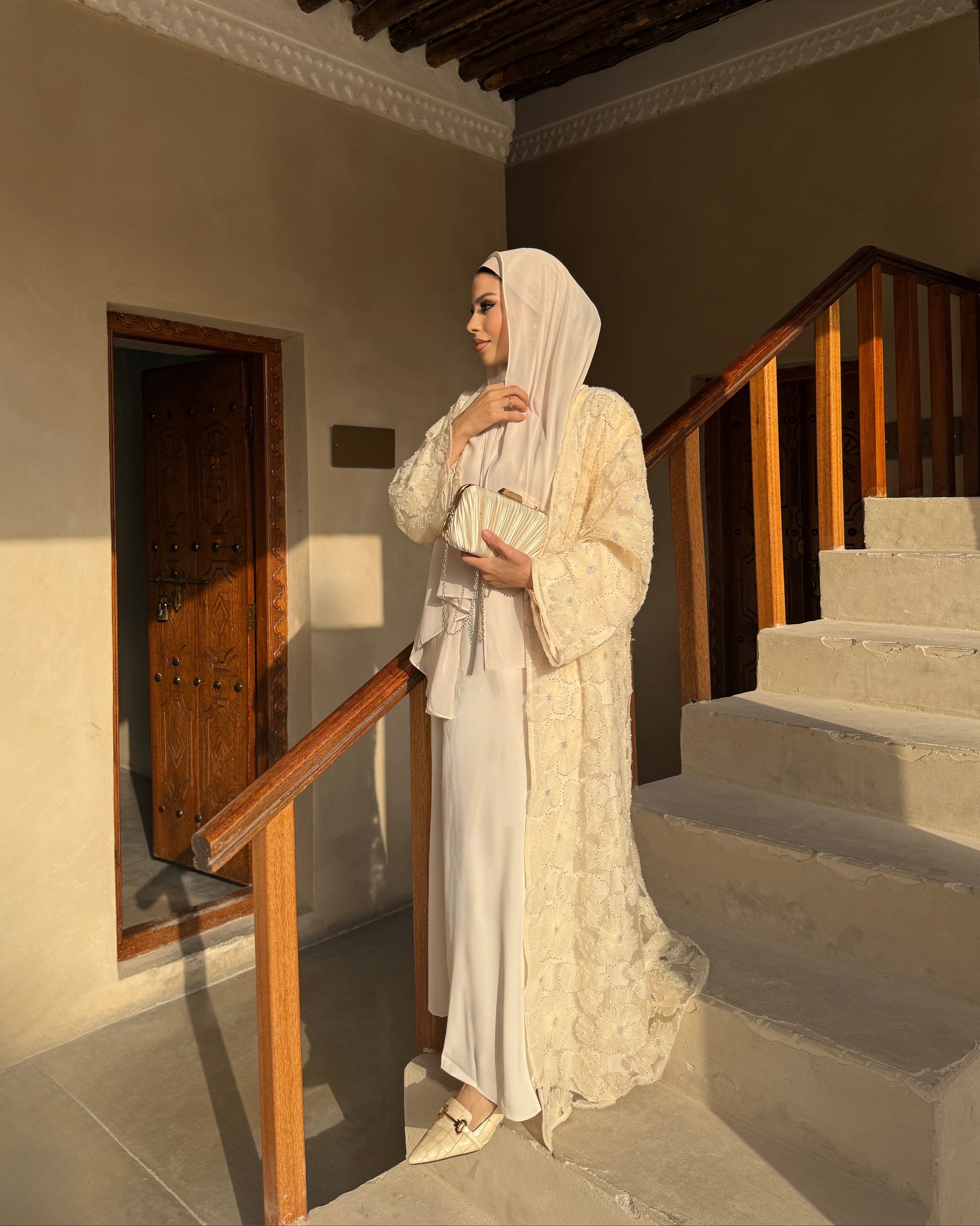 Woman in a Sumayya Yellow Abaya dress standing in a sunlit room with wooden beams and a staircase.