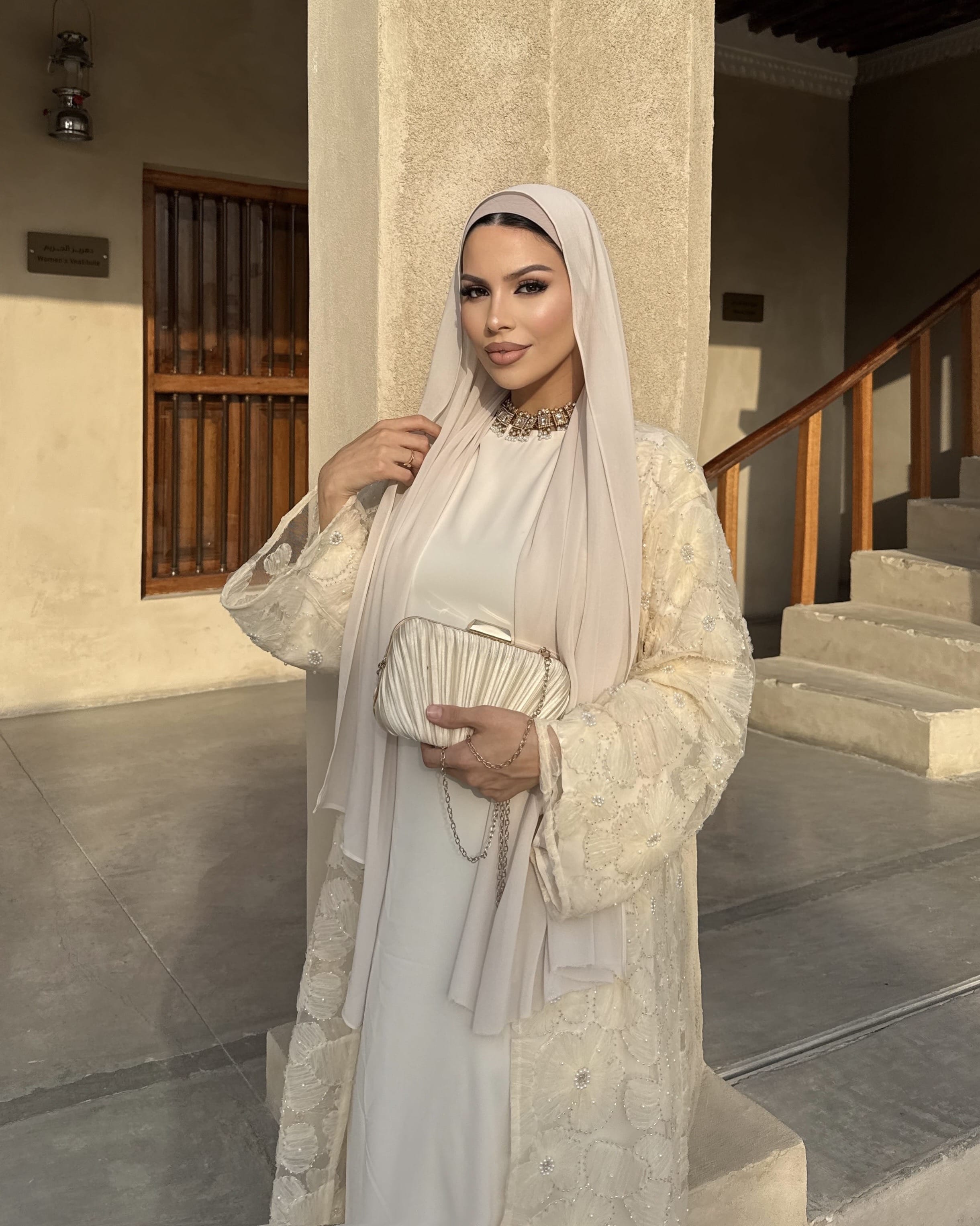 A woman wearing a modest cream-colored outfit with a matching hijab, holding a small pleated clutch bag, standing in an architectural setting with stone steps and columns.